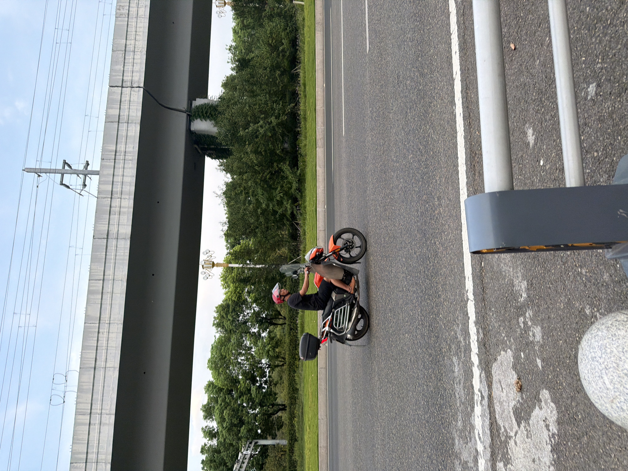 A person is riding an orange motorcycle on an asphalt road in Jiangjin, China. The person is wearing a red helmet and a black shirt. The motorcycle has a black storage box on the back. In the background, there is a raised concrete overpass with electrical wires. Trees and vegetation line the side of the road. The sky is overcast. A white line is painted on the road. In the foreground, there is a gray metal barrier and a rounded gray object.