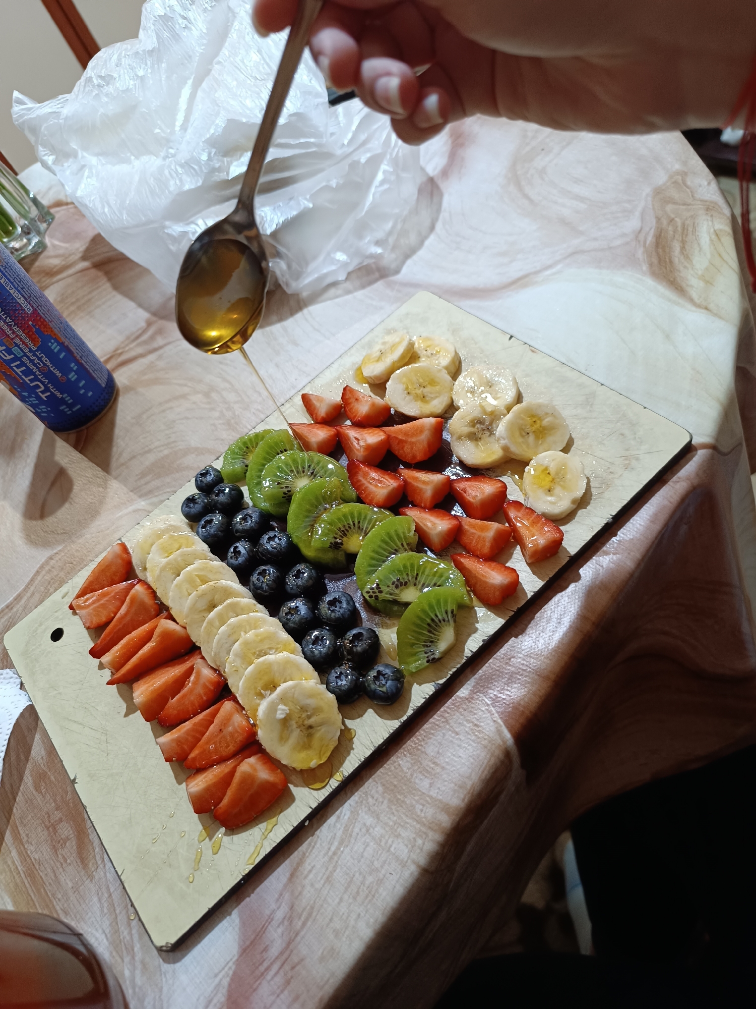 A still life photograph captures a wooden cutting board arranged with rows of sliced fruits: strawberries, bananas, blueberries, and kiwi. A metal spoon containing a clear, golden liquid, possibly honey, is positioned above the fruit, with droplets visible on the fruit and board. The arrangement sits on a surface patterned with an abstract, possibly marble-like design. A hand is visible near the spoon. A blue can is partially visible in the upper left, labelled "TUTTI FRUTTI." The location is not discernible from the image.