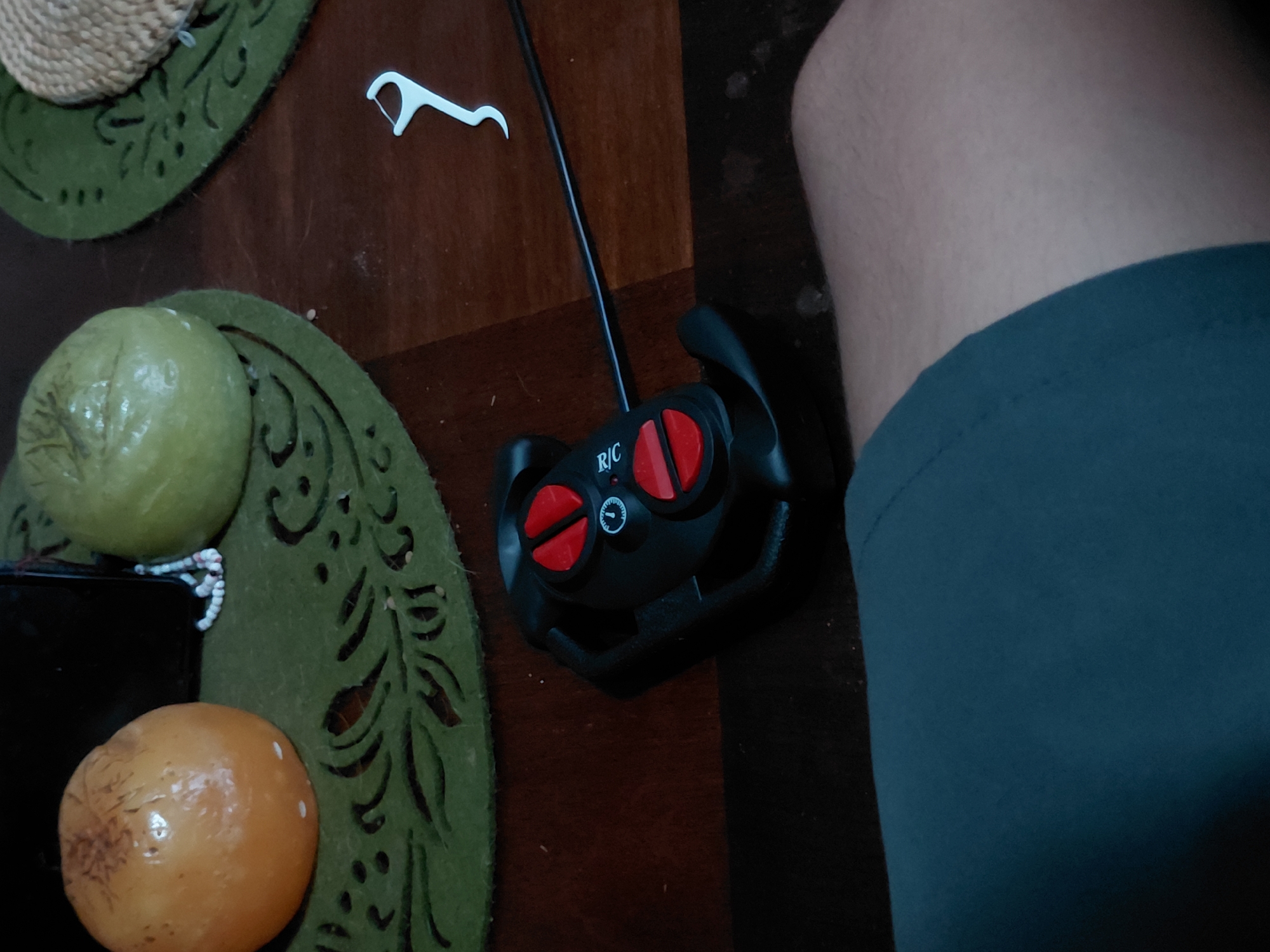 The image depicts a dark brown wooden table surface on which several items are placed. In the upper left corner is a straw hat resting on a green decorative placemat with intricate cutout designs. Next to the hat, a white dental floss pick is visible. Below the hat, on a similar green placemat, are two round objects: one green and the other orange.
A black remote control with a cable is situated near the center of the table, featuring red buttons and the letters "R/C" printed on it. Adjacent to the remote control, a person's leg, covered in gray fabric, is visible.
The image lacks any specific details that would allow for determination of the location of Santa Ana, Venezuela.