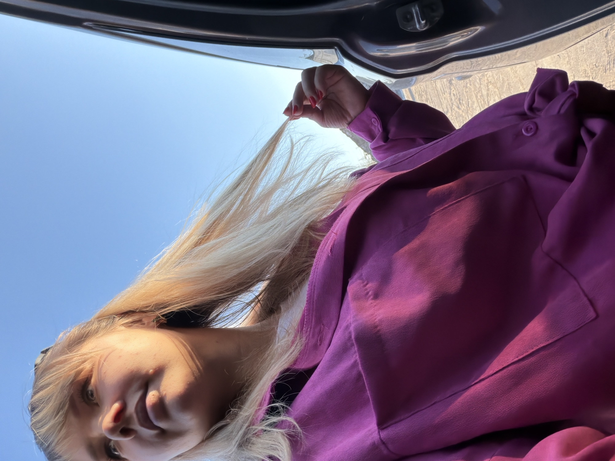A low-angle shot depicts a woman holding a strand of blonde hair in her right hand. She wears a magenta or purple shirt. The background features a bright blue sky and a portion of a car hatch. The location is unknown and there is no way to prove with certainty it is in Chaikovsky, Russia.