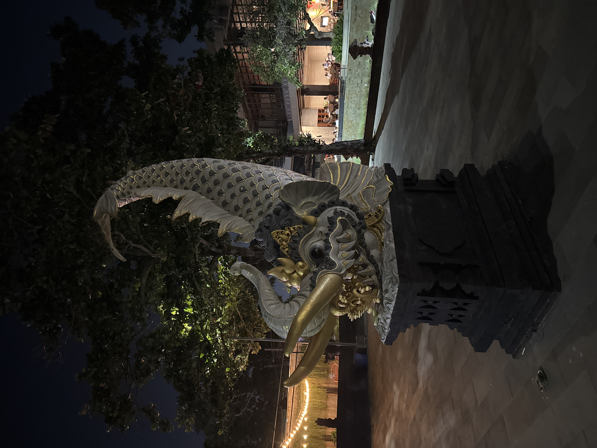 This image captures a nighttime view of a large, ornate statue of a dragon or mythological creature in Kuta, Indonesia. The statue is positioned in an open area paved with patterned bricks, with trees and a building in the background. The statue itself is detailed, featuring scales, large golden horns, and intricate facial features, all illuminated by subtle lighting that casts shadows. Behind the statue, string lights hang across a courtyard, and a multi-story building with open-air seating and diners can be seen. The environment suggests a public space or dining area, likely a restaurant or resort, common in the tourist areas of Kuta. The lighting, time of day, and the statue's style point to an Asian cultural influence, specifically Balinese art.