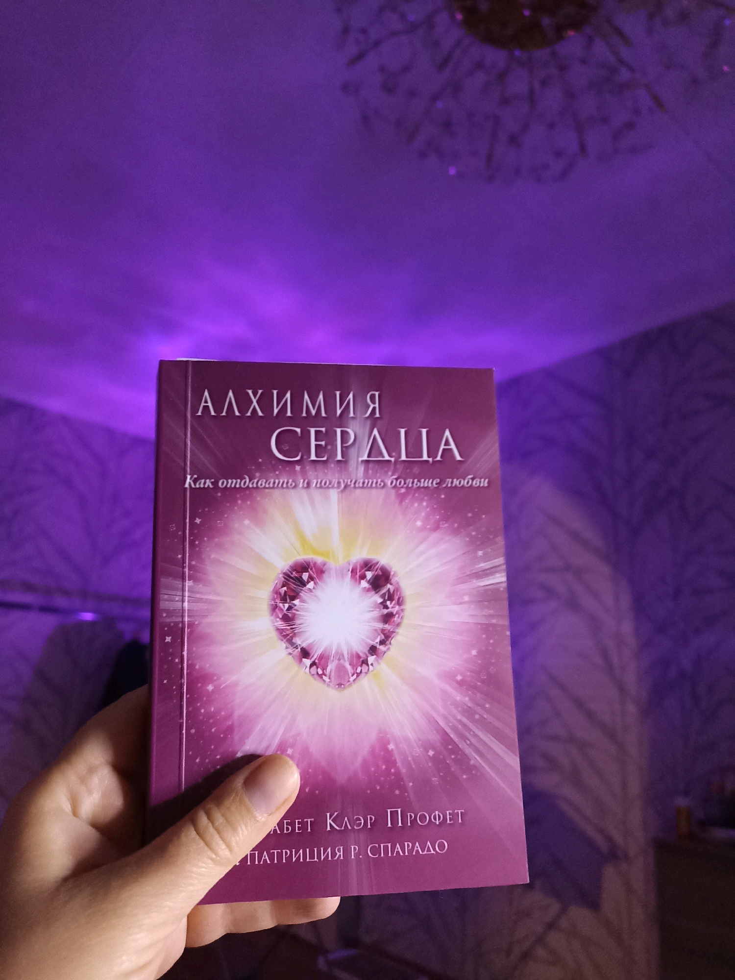 A person's hand is holding a book with a purple cover. The book has the title "АЛХИМИЯ СЕРДЦА" in large white Cyrillic letters. Below that, in smaller white letters, it says "Как отдавать и получать больше любви" (How to give and receive more love). The cover features a stylized pink heart made of gemstones, emanating light. The bottom of the book shows text in smaller Cyrillic letters, including "АБЕТ КЛЭР ПРОФЕТ" and "ПАТРИЦИЯ Р. СПАРАДО". The background is dimly lit and appears to be a room with purple ambient lighting, possibly from a lamp or light fixture, casting shadows on a textured wall. The image does not contain information about the location in Novosibirsk, Russia.
