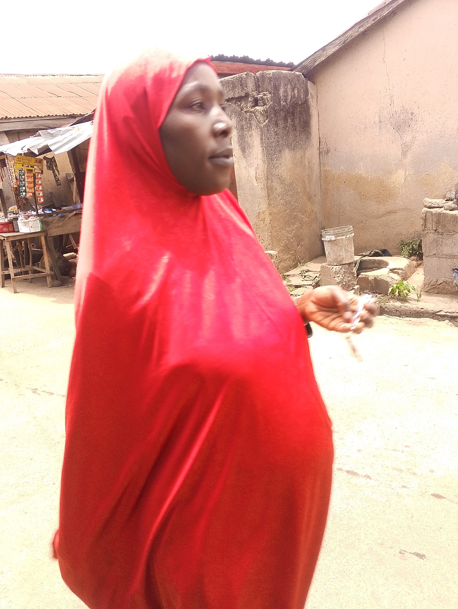 The image depicts a person wearing a red hijab walking on a paved surface. The person appears to be outdoors, possibly on a street. In the background, there are buildings, a shop with goods displayed, and general street debris. The location is presumed to be in Jos, Nigeria, based on the prompt's request. The person is holding a small object in their right hand. The scene is brightly lit.