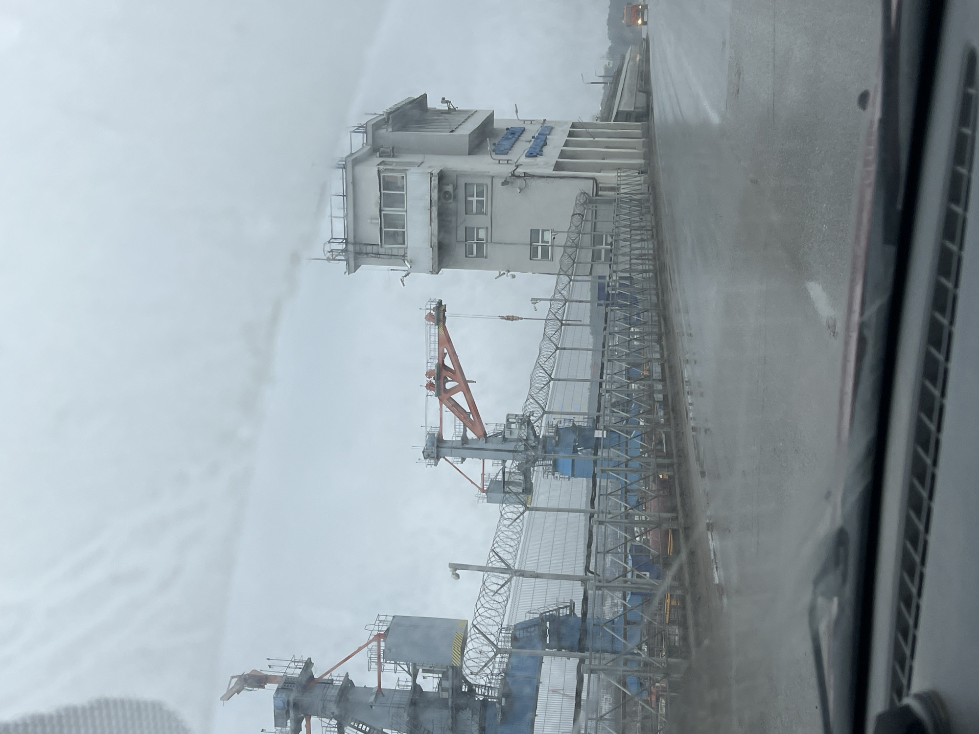 The image shows a view from inside a vehicle, likely a car, looking out towards an industrial area. The weather appears to be overcast and possibly rainy, with a grey sky.
The dominant feature is a tall, industrial building with multiple levels and windows. There appears to be some signage on the building's facade, although the exact text is not clearly legible. A crane with an orange jib is visible in front of the building.
In the foreground, there is a structure of metal scaffolding and barbed wire. This structure runs parallel to the building, and it may be part of a security perimeter or some other form of industrial infrastructure.
The foreground also shows part of the interior of the car, with a section of the dashboard and door visible. The overall impression is of an industrial site viewed through the window of a vehicle in cloudy weather. Based on the prompt, this is likely located in Chaikovsky, Russia.