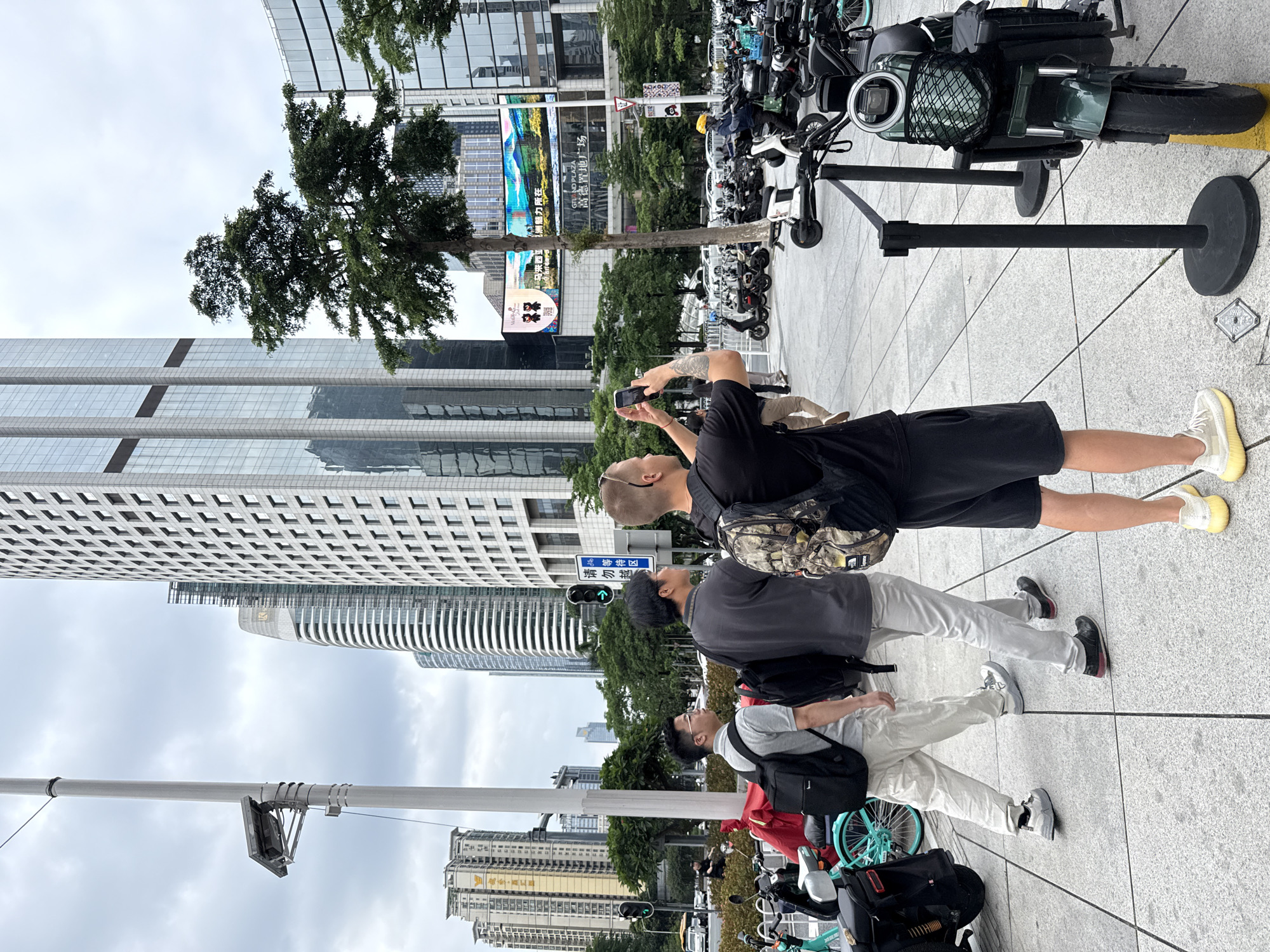 A group of three men are walking on a paved plaza in Tianhe District, China, with a backdrop of modern high-rise buildings and trees. The man in the foreground, on the right, is wearing a black t-shirt, black shorts, and yellow sneakers, and he is taking a photo with his phone, holding it with both hands. He has tattoos on his left arm. To his left, a man in a dark grey t-shirt and light grey pants, wearing a camouflage backpack, walks ahead. The third man, to the far left, is wearing a grey t-shirt, light beige pants, and grey sneakers, also carrying a black backpack. In the background, a large LED screen displays an advertisement with images of mountains and Chinese text. Several electric scooters and bicycles are parked on the right side of the plaza, with a green scooter prominently featured in the foreground. Traffic lights and street signs are visible, indicating an urban environment. The sky is overcast.