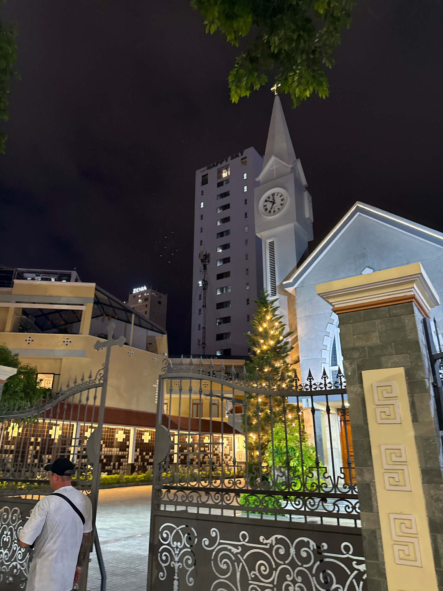 The image depicts a night scene in Nha Trang, Vietnam, focusing on a church and surrounding buildings. In the foreground, a man wearing a white t-shirt and a black cap stands with his back to the camera, facing an ornate metal gate. Beyond the gate, a pathway is visible, leading towards illuminated buildings with intricate patterns on their facades.
To the right of the gate, a tall, gray stone pillar with decorative yellow squares is prominent. Further into the scene, a decorated Christmas tree, adorned with numerous small lights, stands in front of the church. The church itself is light blue with white trim and features a prominent clock tower with a visible clock face. A golden cross tops the church's steeple. Behind the church, a tall, modern building with many illuminated windows stands out against the dark night sky. Another building, labeled "ZENIA," is visible between the church and the taller modern structure. The sky is dark and cloudy, with some faint lights scattered across it.