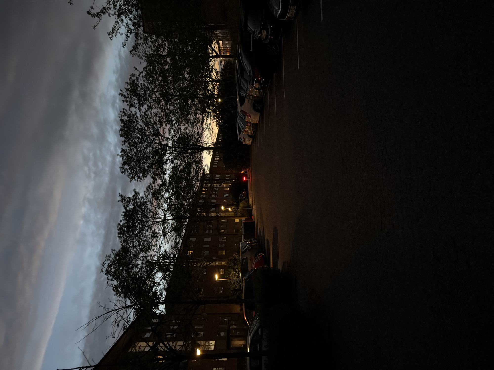 The image depicts a dark, overcast scene in what appears to be a parking lot adjacent to a building in Horsens, Denmark. The sky is filled with heavy clouds, with a hint of light breaking through near the horizon. Silhouetted trees stand between the viewer and the building. Cars are parked in the lot, with headlights or taillights visible on some. Streetlights illuminate the scene, casting pools of light on the pavement. The building has multiple lit windows.
