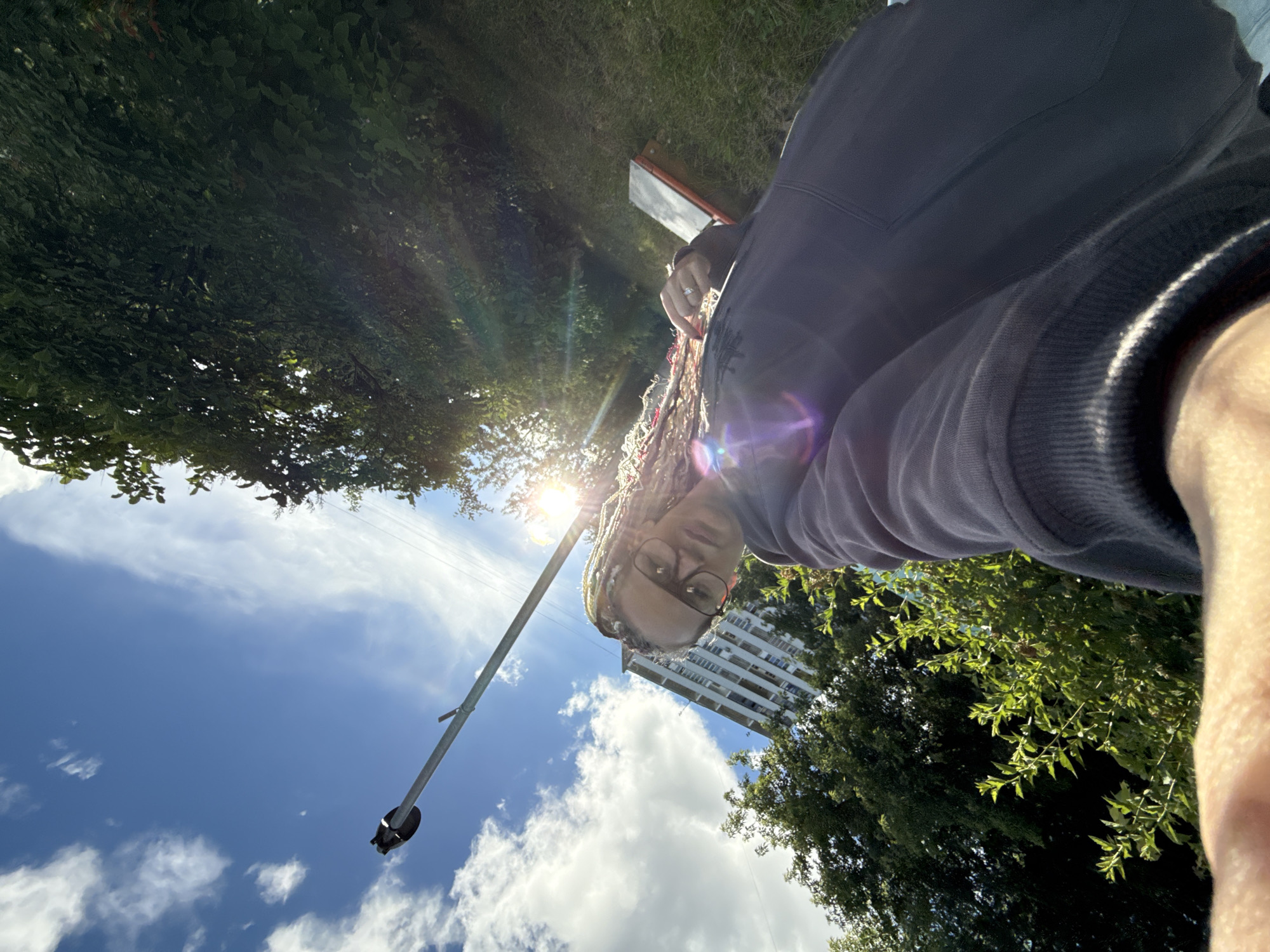 A low-angle selfie shows a person with dreadlocks and glasses against a bright blue sky with scattered clouds. The person is wearing a dark gray hooded sweatshirt. Behind them, a tall pole with a light fixture on top extends upwards, and a multi-story apartment building is visible in the background. Lush green trees and foliage surround the scene, with sunlight streaming through the leaves, creating lens flares. The ground is covered in grass. The image does not contain any information that could identify the location as Moscow, Russia.