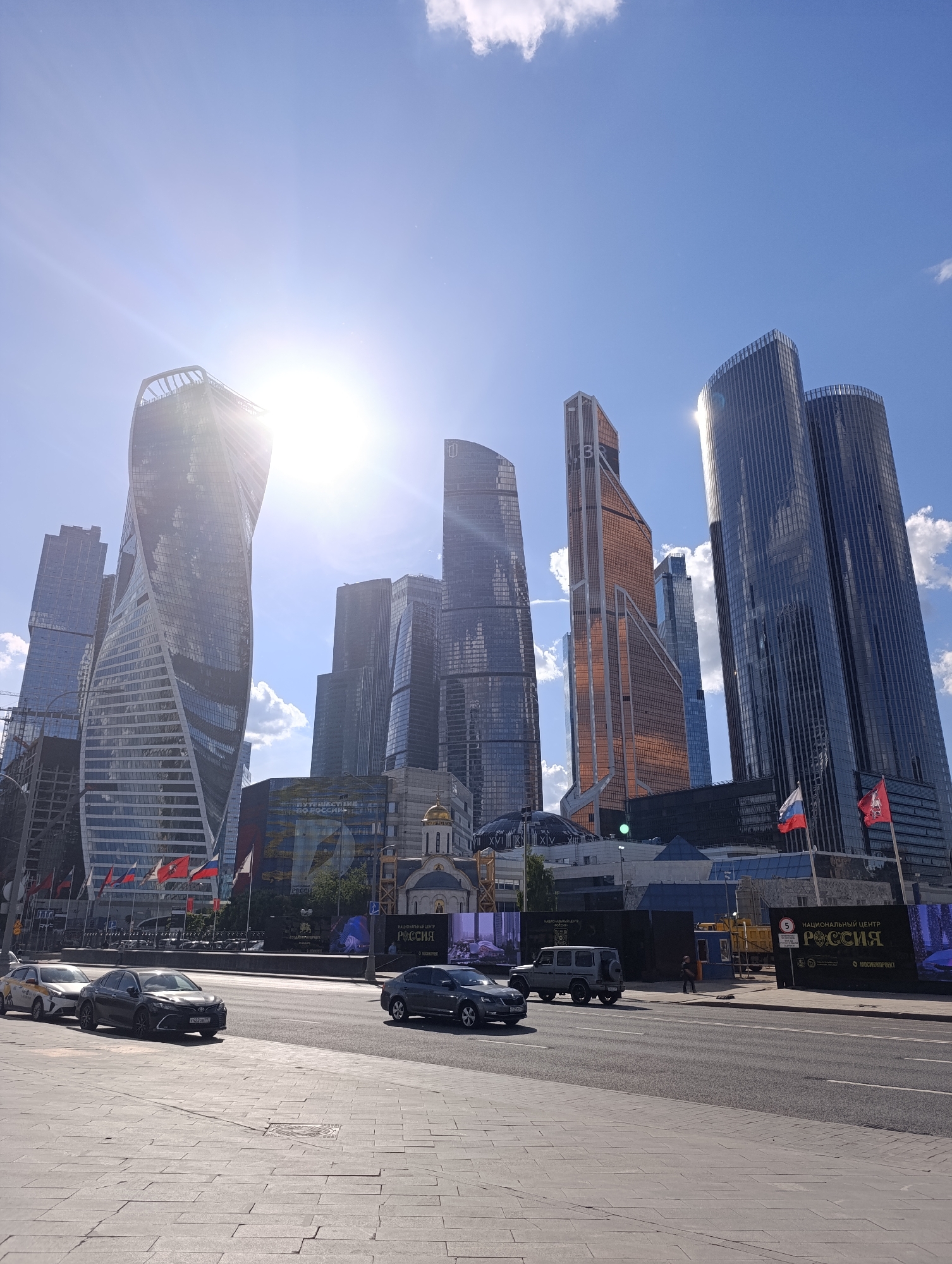 This image captures a bright, sunny day in a modern city, likely Moscow, Russia. The scene is dominated by a cluster of tall, contemporary skyscrapers with glass and steel facades. On the far left, the distinct helical design of the Evolution Tower is prominent, its surface reflecting the intense sunlight. To its right, various other high-rise buildings, including one with a bronze hue and two darker, cylindrical towers, pierce the clear blue sky, which features scattered white clouds. The sun is positioned high, creating a strong lens flare in the upper left quadrant of the image.
In the mid-ground, a wide asphalt road stretches across the frame, with several vehicles present. A white and yellow Yandex Taxi is parked on the left side, followed by a dark sedan. Further down the road, another dark sedan and a grey Mercedes G-Wagen SUV are visible. A single person can be seen walking on the far side of the road near the SUV.
At the base of the skyscrapers, several ground-level structures are visible, including a small, traditional-style building with a golden dome. Adjacent to it, flagpoles display Russian flags and possibly local Moscow flags. Dark construction barriers or large banners line the roadside, featuring Russian text. One banner reads "ПУТЕШЕСТВИЙ ПО РОССИИ" (Journeys across Russia), and another proclaims "НАЦИОНАЛЬНЫЙ ЦЕНТР РОССИЯ" (National Center Russia) with "МОСЖИЛПРОЕКТ" (Moscow Housing Project) below. A speed limit sign indicating "5" is also visible. The foreground features a light-colored paved sidewalk. The bright conditions and visible text strongly suggest a location within Moscow, likely the Moscow International Business Center (Moscow City).