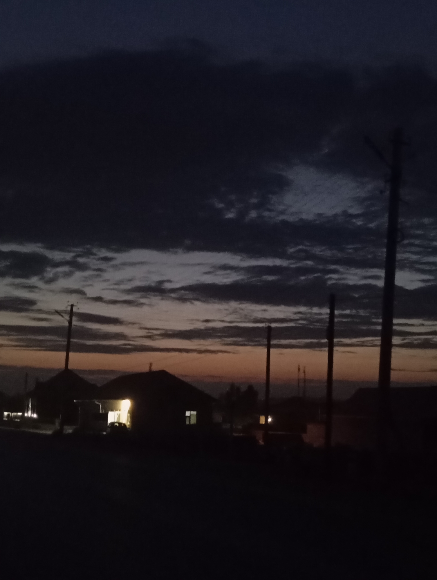 The image depicts a dark, twilight scene in what is identified as the city of Kızıl-Bel, Kyrgyzstan. Silhouetted against a dusky sky are several residential structures, the roofs of which are dimly visible. One house in the center of the frame is illuminated from within, with a single light source casting a yellow glow. A car is parked in front of this illuminated house. Power lines and poles are prominent in the foreground and midground, extending upwards into the cloudy sky. The sky itself is a blend of dark blues and grays, with hints of orange and yellow near the horizon, suggesting either sunrise or sunset. The overall atmosphere is serene and quiet, characteristic of a rural or suburban setting at the end of the day.