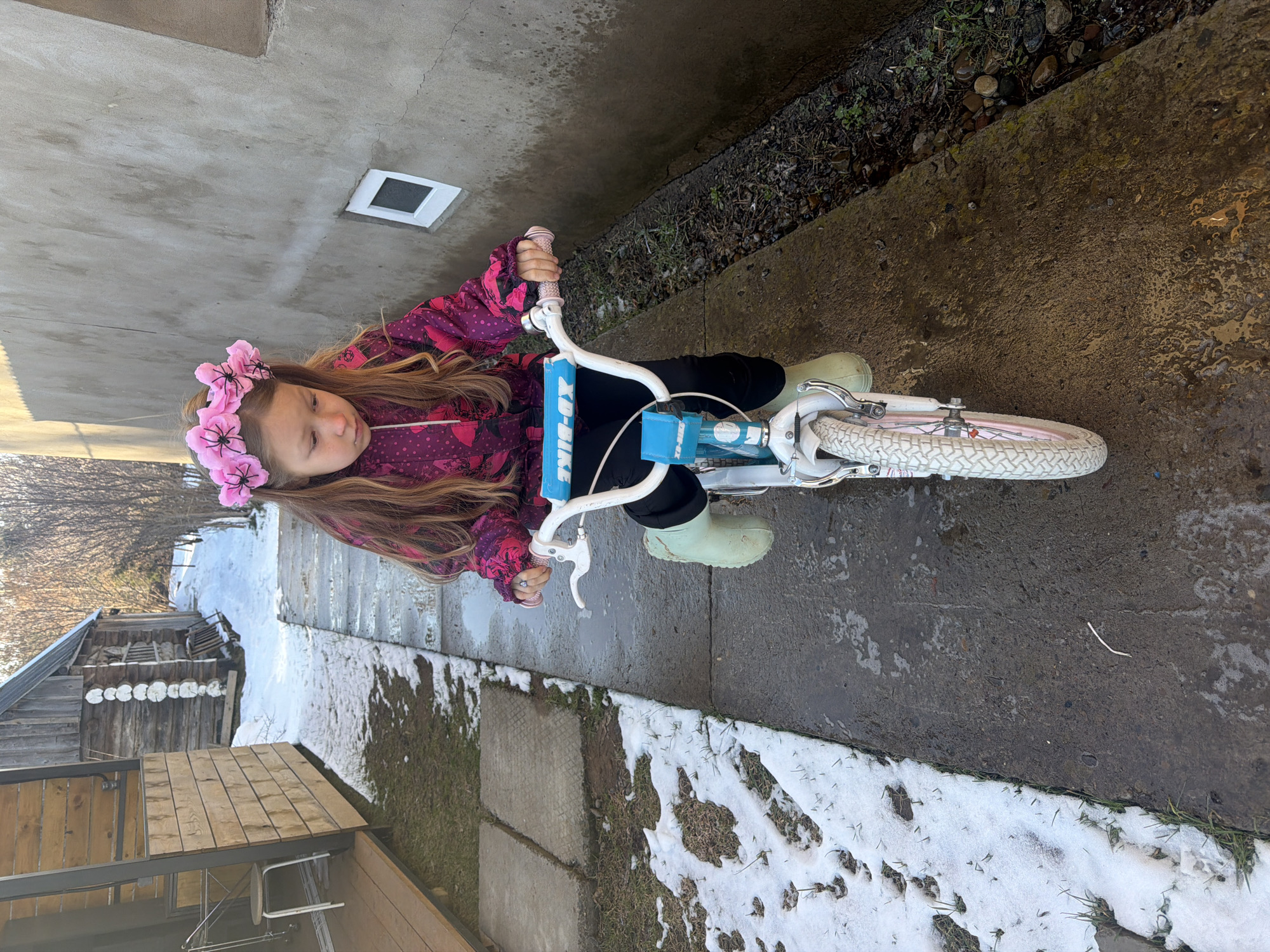 A young girl with long brown hair, adorned with a pink floral headband, sits on a blue and white bicycle outdoors. She is wearing a pink and purple jacket with a heart pattern, black pants, and light green boots. The bicycle is white with pink accents on the wheels. The ground is a mix of concrete and snow-covered grass, with some patches of bare earth visible. A building is partially visible in the background. The location appears to be an urban or suburban residential area.