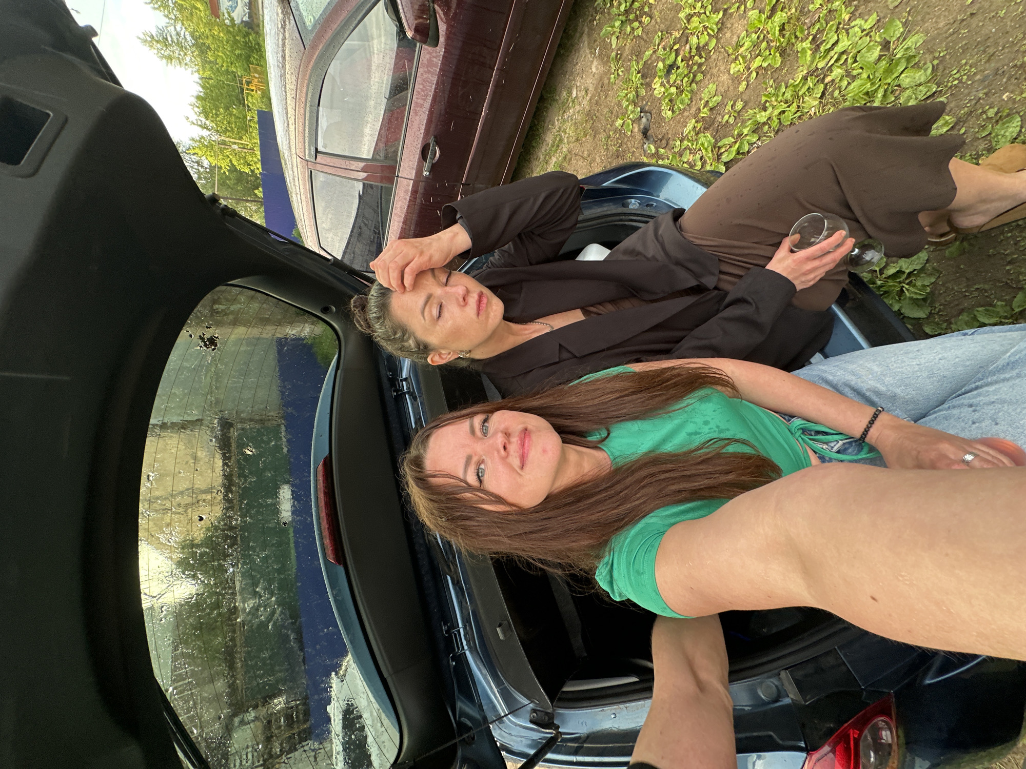 Two women are sitting in the open trunk of a blue car. The woman on the left is smiling at the camera, wearing a green t-shirt and blue jeans. The woman on the right is looking down with her hand on her forehead, wearing a black blazer and a brown skirt. She is holding a wine glass. The car is parked outside, with a maroon car visible in the background. The ground is covered with green plants and there are droplets of water on the car's rear window, suggesting it might be raining or has recently rained. The scene appears to be in a rural or suburban area.