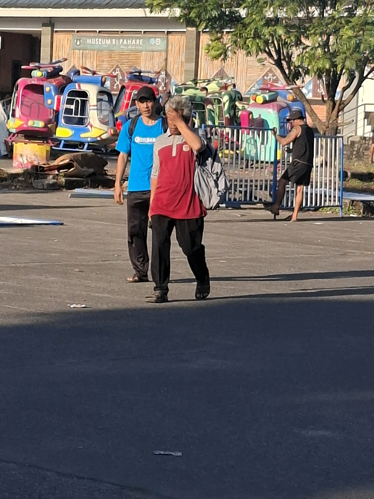 The image depicts an outdoor scene in front of a building with a sign that reads "MUSEUM KI PAHARE." Several colorful children's amusement rides, resembling helicopters and tanks, are visible. Two men are walking in the foreground. The man on the left wears a blue t-shirt and black pants, with a black cap and a backpack. The man on the right wears a red and grey shirt, black pants, and has his hand over his face. In the background, a third individual, wearing a black tank top and shorts, is seen near a fence, with one leg raised as if dancing or posing. The ground is paved asphalt, and a tree with green foliage stands to the right of the frame. The overall setting appears to be a public space, possibly an amusement area or park, adjacent to the museum. The lighting suggests it is daytime, with shadows cast on the ground.