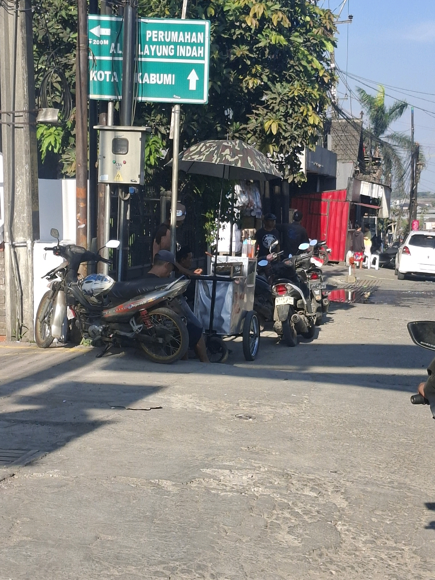 In Lembursawah, Indonesia, a street scene unfolds under a clear sky. A traffic sign indicates "PERUMAHAN LAYUNG INDAH" (Layung Indah Housing) with an upward arrow, and "F 200M AL KOTA KABUMI" (200m to AL City Kabumi). A utility box is mounted on a pole.
In the foreground, a silver motorcycle is parked, and another motorcycle is parked next to it. A small cart with a camouflage umbrella is also present, and people are gathered around it. In the background, several buildings are visible, including one with red siding. A white car is parked on the right side of the street. The street itself is paved and shows some signs of wear. The environment appears to be a typical urban or semi-urban setting in Indonesia.