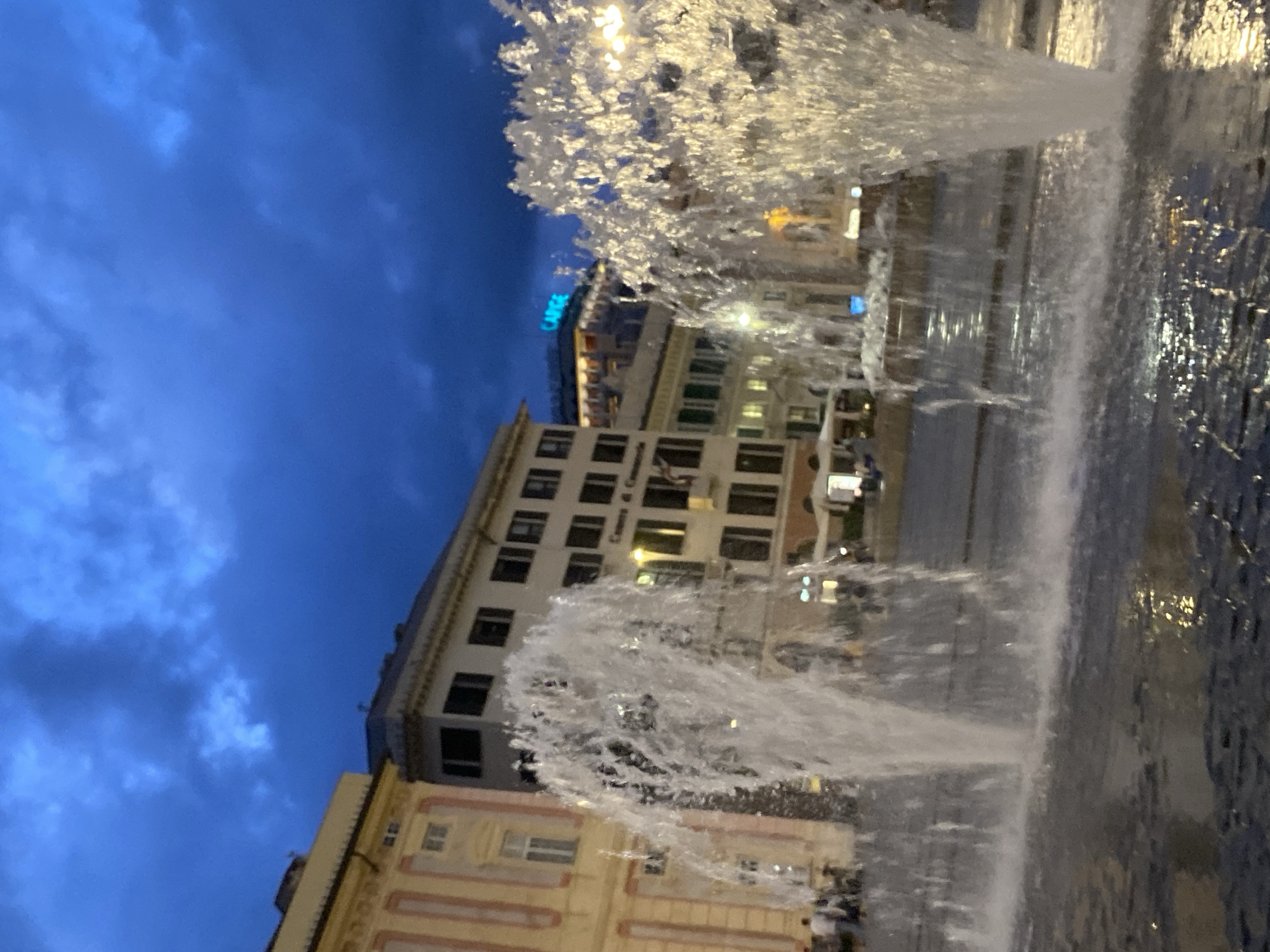 This is a nighttime, low-angle shot of a large public fountain in Piazza de Ferrari, Genoa, Italy. The sky is a deep, cloudy blue, suggesting twilight or impending rain. Two large water jets dominate the foreground, one on the left and one on the right, spraying water into the air and creating a frothy, dynamic effect. The water falls back into a dark, wet paved area, reflecting streetlights.
In the background, several buildings are visible. On the left, a pale yellow building with ornate details and arched windows stands. Adjacent to it is a taller, light-colored building with multiple dark rectangular windows. Further back, a modern building with a bright blue sign reading "CARIGE" is illuminated. In the middle ground, other buildings with illuminated windows and awnings are visible, as well as a few scattered streetlights. Several people are visible in the mid-ground, some walking, and others appearing to be seated at outdoor cafes. The overall impression is of a lively public square at dusk.