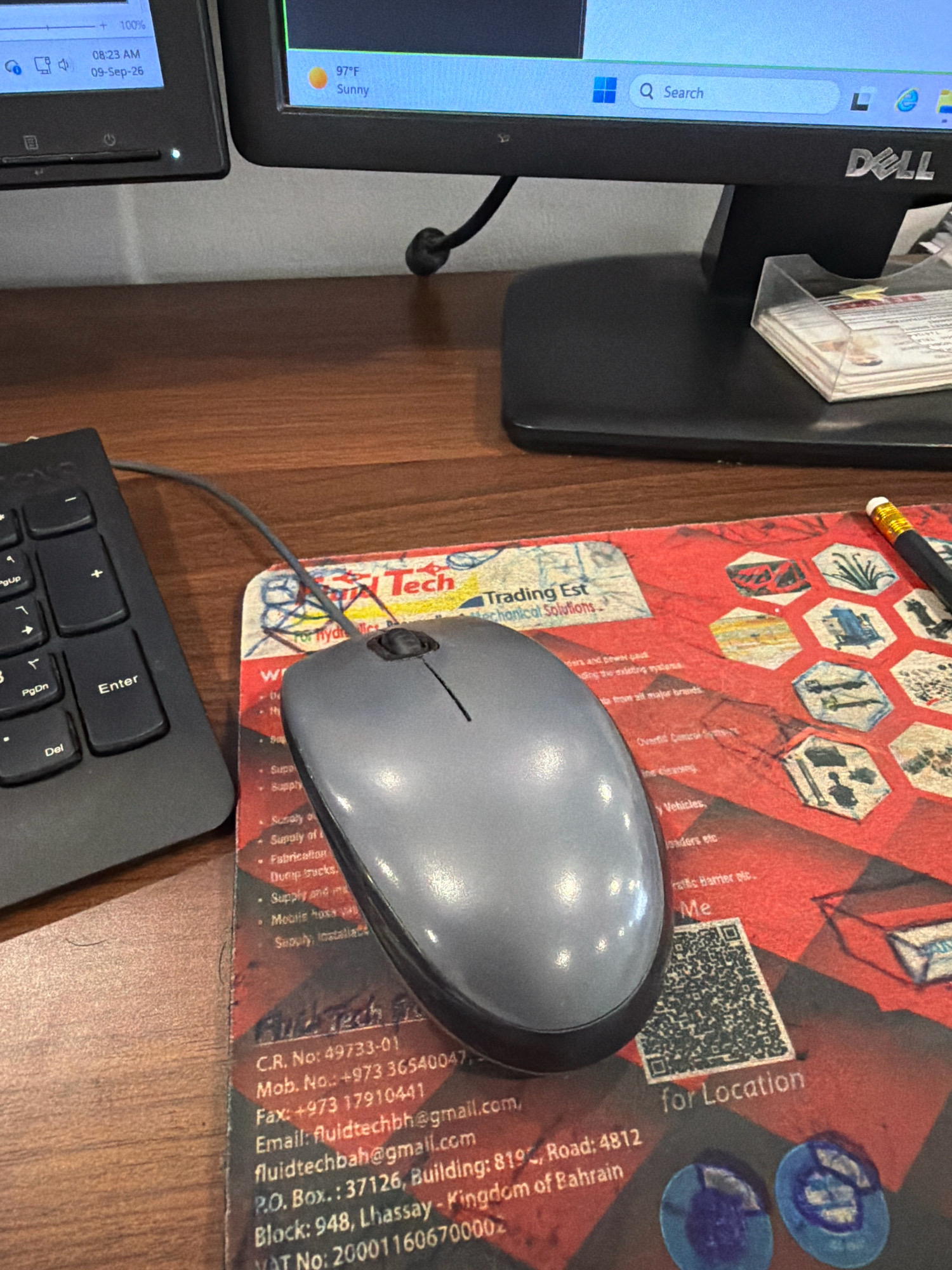 The image shows a desk with two computer monitors, a keyboard, and a mouse on a mousepad. The mousepad has a red background with various hexagonal images and text. The text on the mousepad includes "Trading Est Mechanical Solutions" and contact information for "Fluid Tech". The contact information includes a CR number, mobile number, fax number, email address, P.O. Box, and address. The address is "Block: 948, Lhassay - Kingdom of Bahrain". This indicates the location is in the Kingdom of Bahrain, with Lhassay being a specific area within the country. While Riffa is a city in Bahrain, the provided address does not specifically mention Riffa. One monitor displays the time as 08:23 AM on 09-Sep-26, and the other displays the temperature as 97°F and "Sunny". A pencil is visible on the right side of the mousepad. The overall environment appears to be an office or workspace.