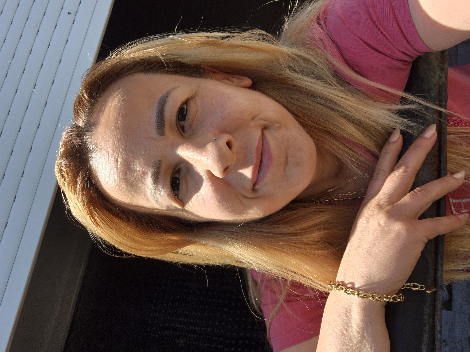 A medium shot shows a fair-skinned woman with light blonde hair, wearing a pink shirt and a gold bracelet. She is leaning on a dark metal bar, her right hand resting on it. The woman has a slight smile, with a nose ring visible. Sunlight casts shadows on her face. The background is a dark, textured surface and a white, slatted structure.