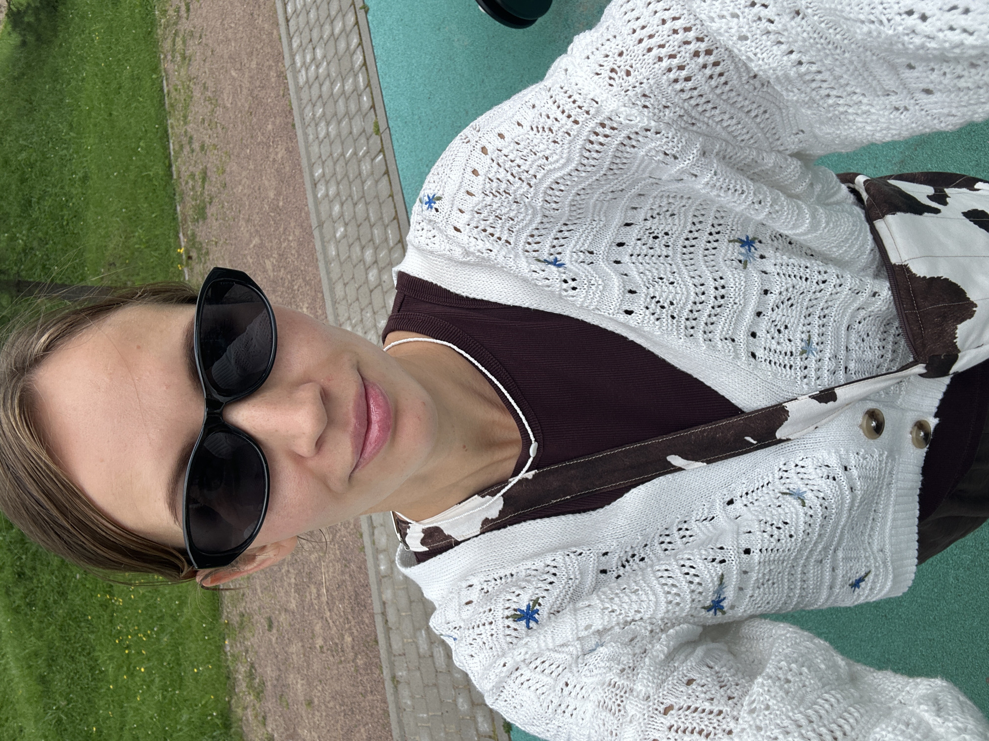 A young adult woman with light brown hair, wearing sunglasses, a white knitted cardigan with blue floral embroidery, a dark purple ribbed top, and a purse with a cow-print strap is standing outdoors. She is looking at the camera and smiling. The background consists of grass, gravel, and paving stones, suggesting an outdoor public space such as a park or playground in Vsevolozhsk, Russia.