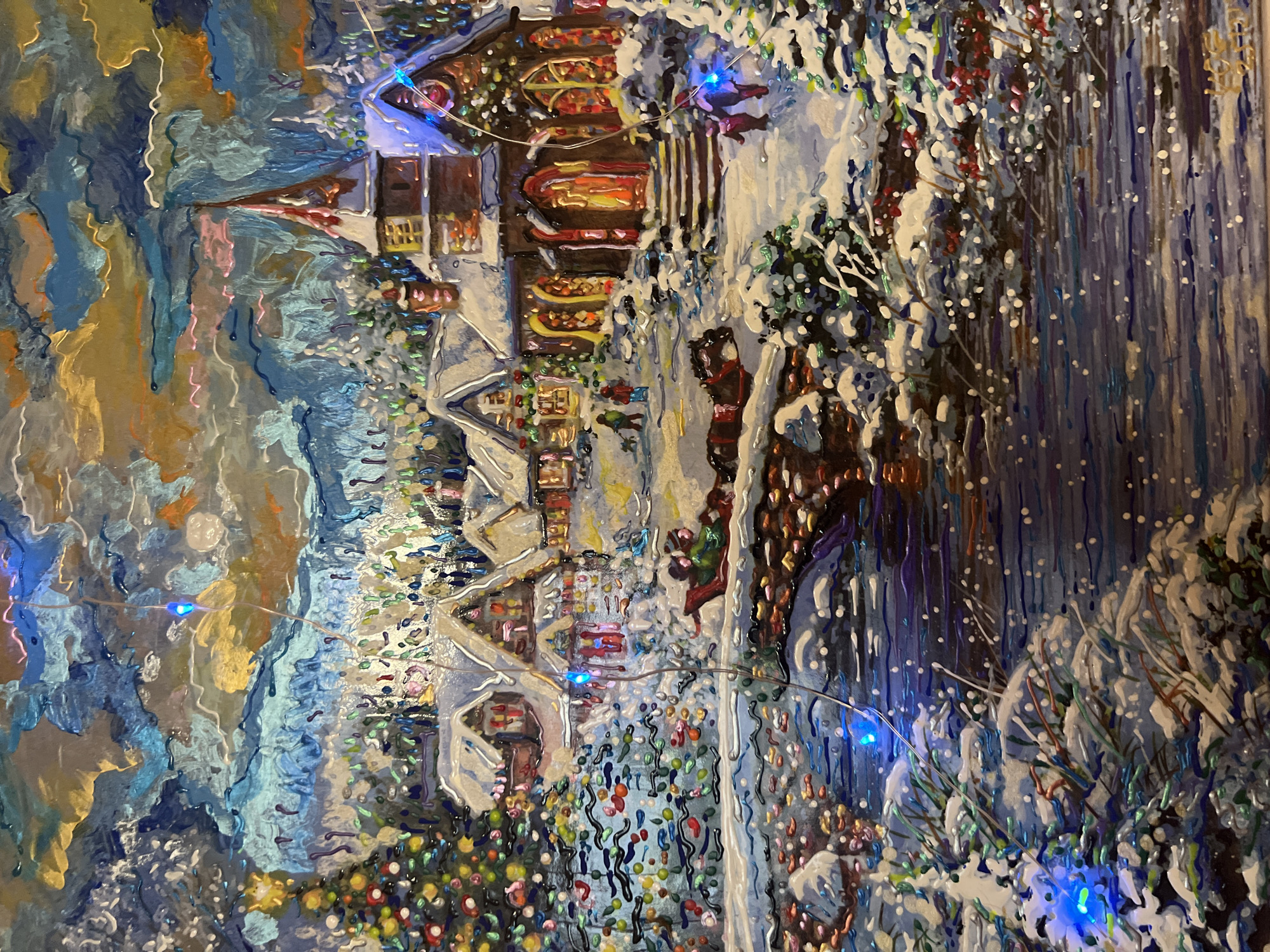 The image is a painting of a snowy Christmas village scene. A decorated Christmas tree with colorful ornaments and a star on top stands to the left. In the background, there are several snow-covered houses and a church with a steeple, all illuminated with warm lights. A bridge arches over a body of water in the foreground, with a horse-drawn sleigh crossing it. Several figures are depicted walking in the snow, some with dogs. The sky is filled with swirling clouds in shades of blue, grey, and gold, and decorative Christmas lights are woven throughout the scene, adding a festive glow. The painting has a textured, impasto style. There is no indication of the location being Minsk, Belarus.