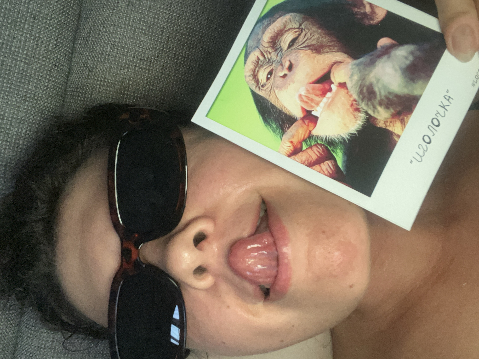 A person lies on a gray fabric surface, possibly a couch, wearing sunglasses and sticking their tongue out. They are holding a Polaroid photograph of a chimpanzee with its mouth open, teeth visible, and someone examining the teeth. There is writing in Cyrillic on the Polaroid, which reads "Иголочка." There is no verifiable information within the image that confirms the location as North Kuta, Indonesia.