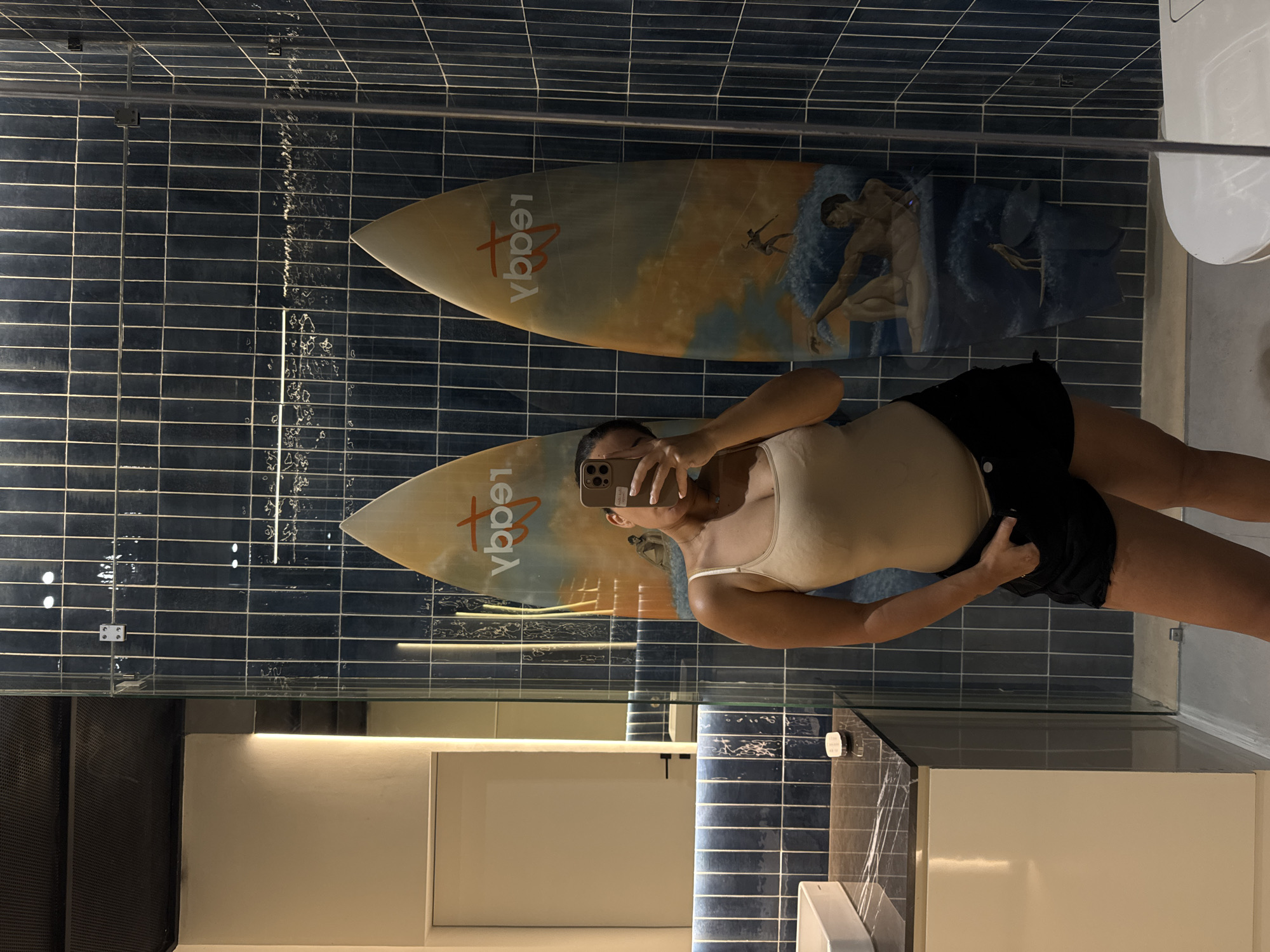 A person is taking a selfie in a bathroom. Two surfboards are mounted horizontally on a blue-tiled wall, one above the other. The surfboards feature an illustration depicting a man and a wave. The bathroom includes a white countertop, a sink, and a clear shower partition. The location is not specified beyond the information already provided.