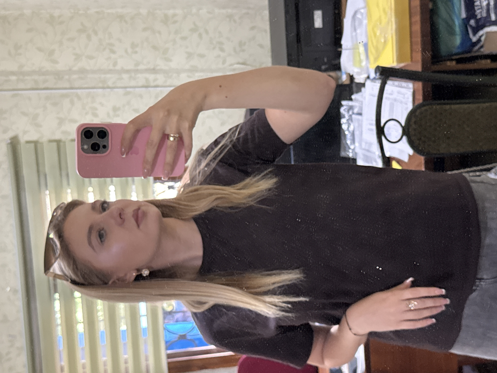 A young Caucasian woman with blonde hair and blue eyes takes a mirror selfie in a room. She is wearing a dark t-shirt, sunglasses perched on her head, and a pearl earring in her left ear. She is also wearing two rings on her right hand, one a gold band and the other a ring with a sparkling design. She holds a pink iPhone in her right hand, which is angled to capture the mirror's reflection. Her nails are painted with a French manicure. The background of the room includes a window with horizontal blinds, a patterned wallpaper, a desk with papers and a printer, and a chair. The scene suggests an indoor setting, likely a home or office.