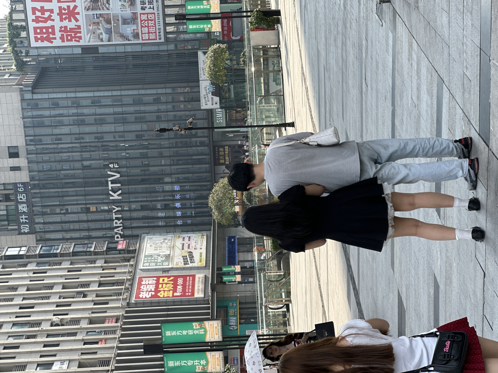 In the Chongqing High-tech Zone, China, a couple stands on a paved city street, facing away from the camera. The man, on the right, wears a light gray t-shirt and light blue jeans, carrying a white purse over his right shoulder. The woman, on the left, is dressed in a black dress with white lace trim at the hem, white socks, and black shoes. She holds a white umbrella with a bear pattern over her left arm. Behind them, large glass-fronted buildings with vertical lines dominate the urban landscape. Advertisements are visible on the building facades, including "PARTY KTV" and Chinese characters for various businesses. The street appears to be a public plaza with a clean, modern design, featuring glass barriers and what seems to be an escalator or stairs leading down. The lighting suggests a sunny day.