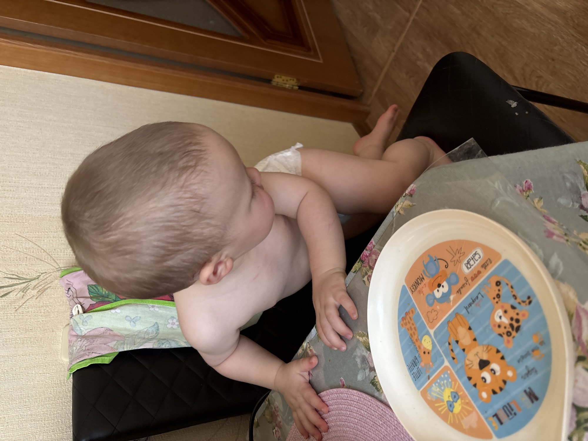 A baby is lying on a black quilted surface, possibly a chair or a mat, with their diaper on. The baby is looking to the right, with their head turned and their eyes focused. Their right arm is bent at the elbow, with their hand resting on a table. A patterned tablecloth is visible underneath the baby and on the table. A white plate with colorful animal illustrations is also on the table. The illustrations include a monkey, a giraffe, a leopard, a tiger, and the sun. Text on the plate includes "cute monkey", "little giraffe", "leopard", "safari friends", and "my little". A pink hat is partially visible next to the plate. The background shows a wooden cabinet and a wall with a textured pattern. The lighting suggests it is daytime. The overall scene appears to be indoors, likely in a home environment. The baby seems relaxed and engaged with something on the table.