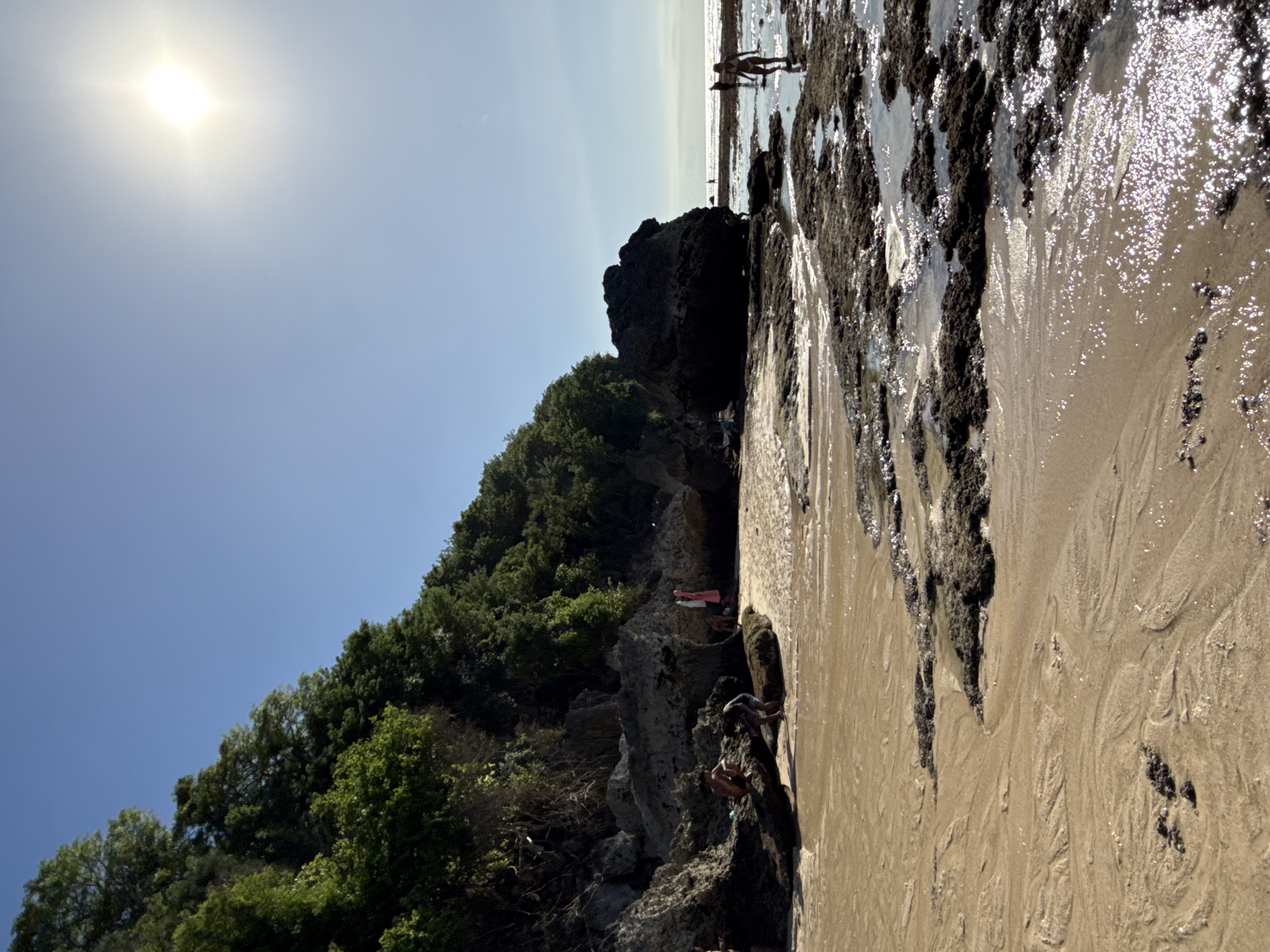The image shows a coastline at Kuta Selatan, Indonesia. The scene is sunlit under a clear blue sky. In the foreground, a beach is visible with exposed rocks and tide pools. Several people are walking along the coastline, both in the shallow water and on the rocks. A rocky cliff covered with dense green vegetation forms a backdrop on the left. The sand on the beach is a light tan color, marked with patterns from the receding tide.
