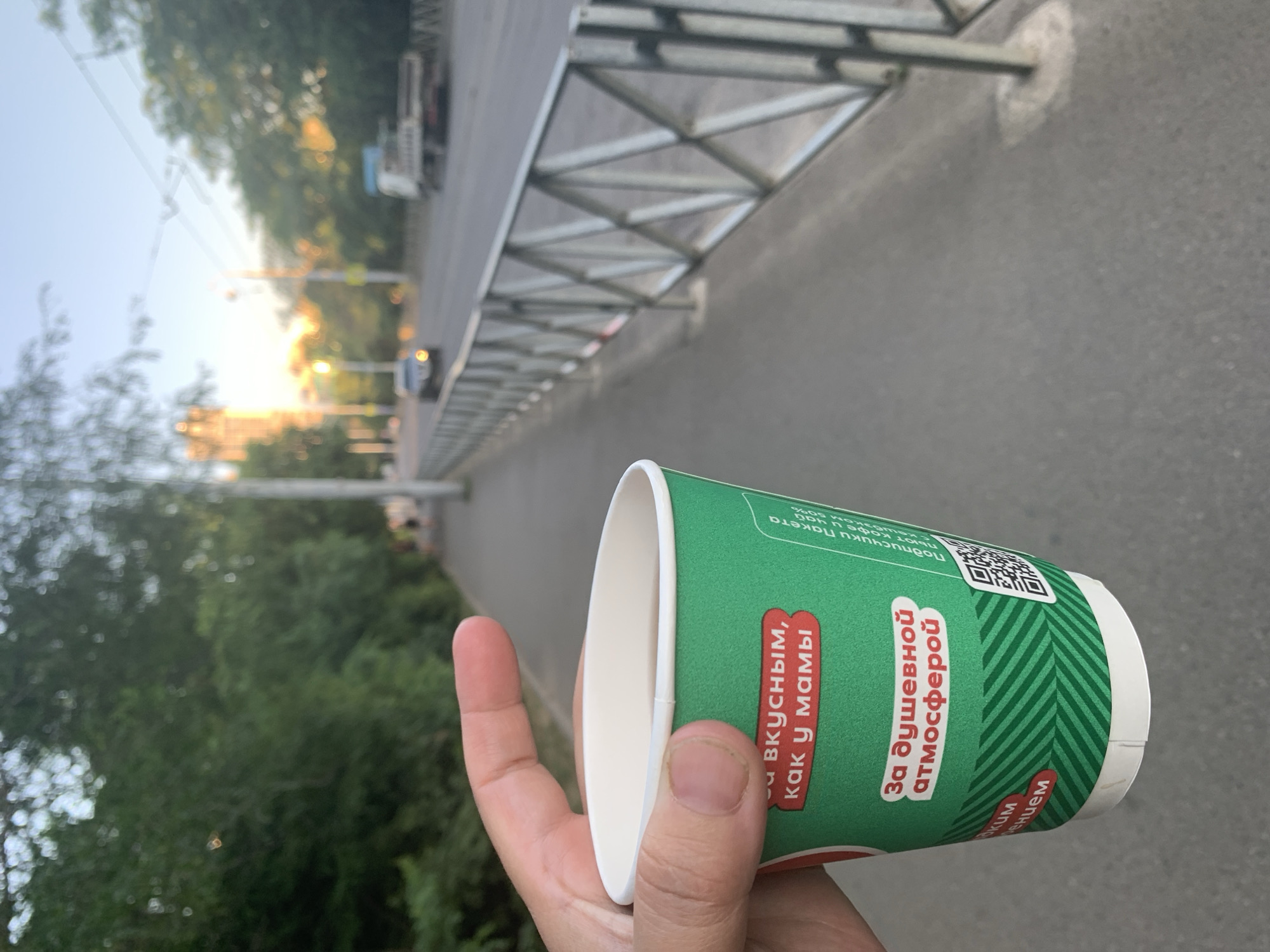 A first-person perspective photo shows a hand holding a green and white paper cup. The cup has text printed on it in Russian, including "Вкусным, как у мамы" (Delicious, like mom's) and "За душевной атмосферой" (For a soulful atmosphere). A QR code is also visible on the cup. The background is blurred, showing an outdoor scene with trees and a grey paved surface that appears to be a sidewalk or road. The lighting suggests it's either early morning or late afternoon, with soft sunlight. The scene appears to be in an urban or suburban environment, possibly a park or along a street. No specific activity is evident other than holding the cup.