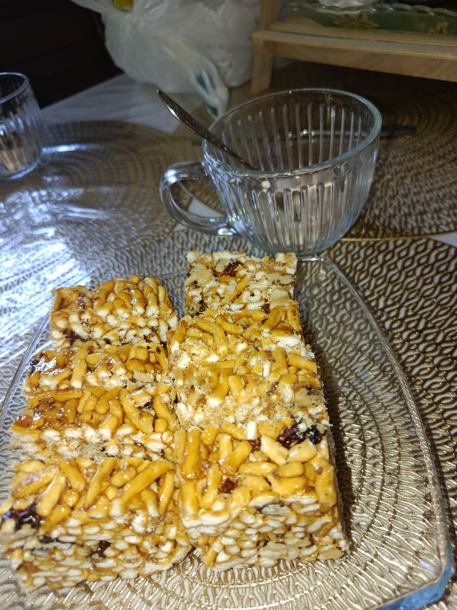A close-up, high-angle shot captures several pieces of sweet, golden-brown confectioneries, likely a type of bar made from puffed rice or similar grains bound together with syrup and possibly nuts or dried fruit, arranged on a clear glass plate. The plate rests on a textured placemat with a repeating geometric pattern in gold and brown tones. To the right of the plate, an empty, ridged glass mug sits on the placemat, with a small silver spoon inside it. In the background, a white plastic bag is visible, and further back, a wooden shelf or furniture piece. The lighting in the scene creates strong shadows and highlights, particularly on the glass surfaces and the placemat. The image appears to be taken indoors, in a domestic setting. The specific location in Tashkent, Uzbekistan, cannot be determined from the visual information provided.