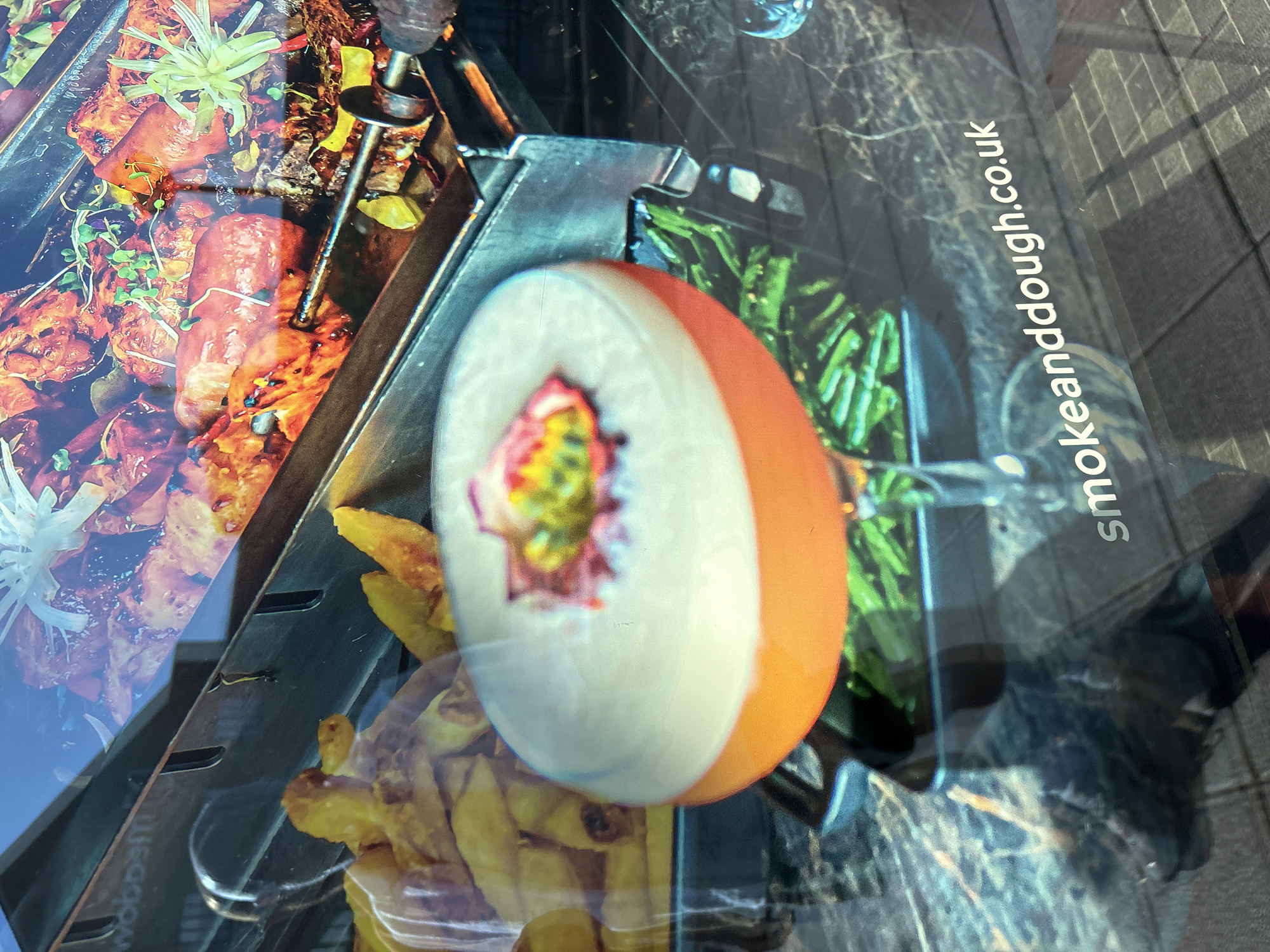 The image shows a close-up view of a cocktail and food displayed behind glass, possibly in a restaurant window. In the background, skewers of grilled meat and fries are visible. In the foreground, a colorful cocktail with a frothy top and garnished with fruit sits in a stemmed glass. The glass is placed on a table with a marble-like pattern. Reflected in the glass, a portion of a website address, "smokeanddough.co.uk," is visible, suggesting the establishment's name is Smoke and Dough. The website address indicates the location is likely within the United Kingdom, and a search reveals that Smoke and Dough has a branch in Chelmsford, United Kingdom. The lighting suggests it might be daytime, with some external light reflecting off the surfaces.