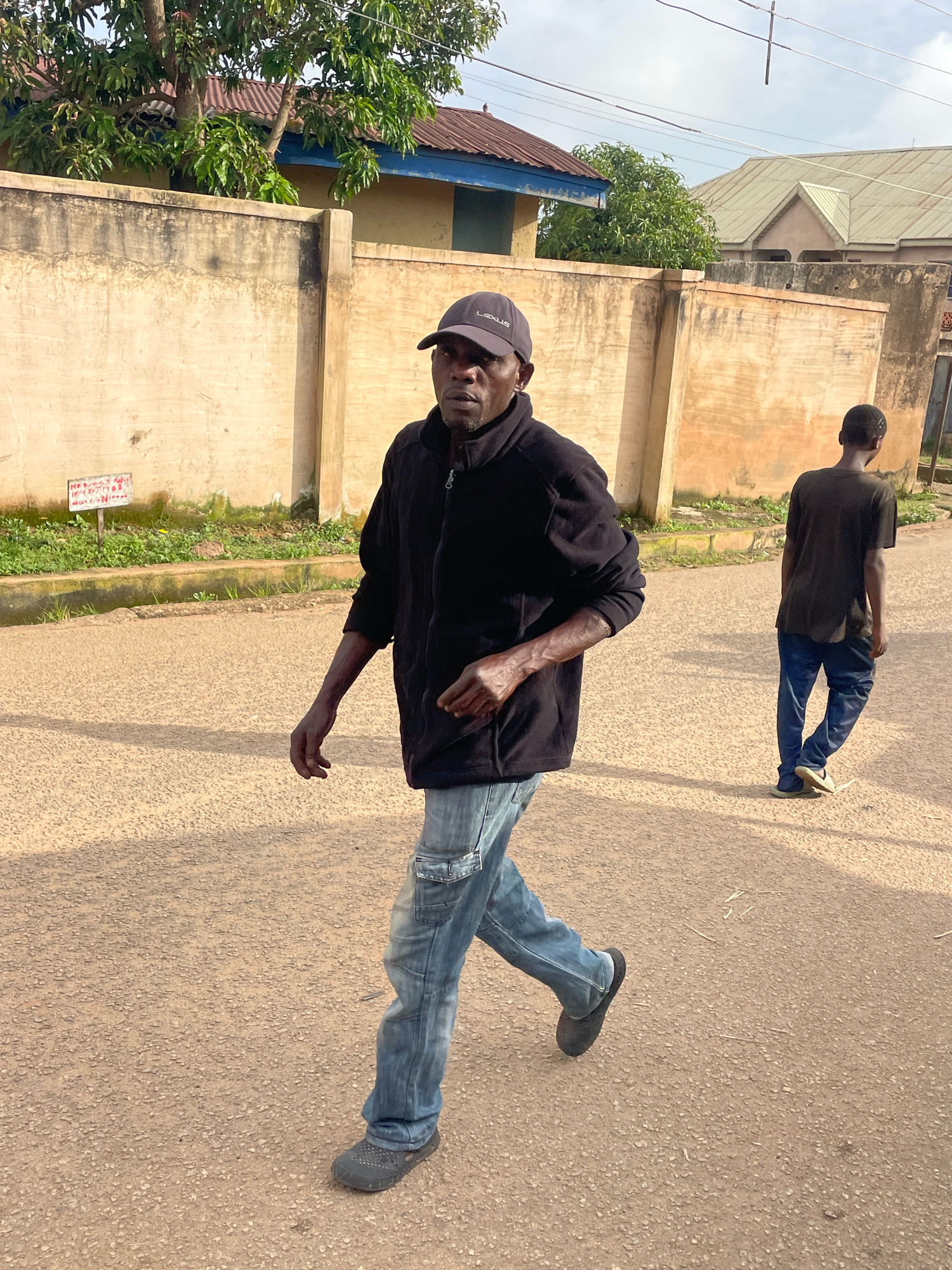 Two men walk along a paved road in Jos, Nigeria. The man in the foreground, wearing a black zip-up jacket, blue jeans, and a dark baseball cap with the word "LEXUS" visible, walks towards the left of the frame. He has his right arm slightly extended and his left arm bent. The second man, wearing a dark t-shirt and blue jeans, walks away from the camera towards the right. A beige wall with some greenery and a small sign is visible behind the man in the foreground. Buildings with corrugated iron roofs are visible in the background. The ground is a light brown, asphalt-like surface with small stones. The sky is visible and appears overcast.