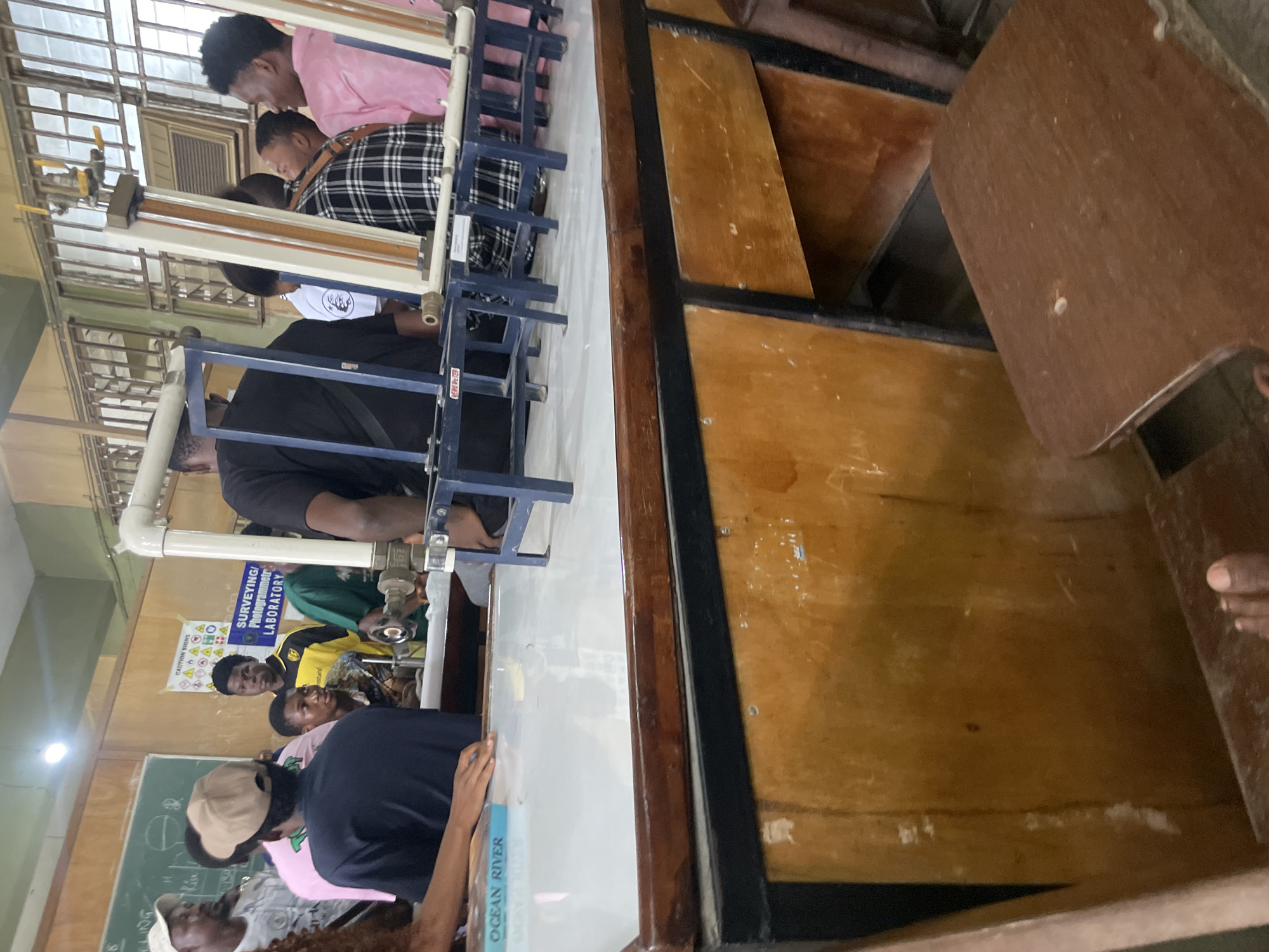 The image depicts an indoor scene, likely a classroom or laboratory, in Ihiagwa, Nigeria. A group of young individuals, predominantly male, are gathered around a table with scientific equipment. The table appears to be a workbench, with a white surface and wooden casing. On the table, there are various pieces of apparatus, including pipes, stands, and what seem to be measuring instruments. Several students are looking at the equipment or engaged in conversation.
In the background, there is a chalkboard with some writing and diagrams. A poster with safety or informational text is also visible on the wall. The overall impression is of an educational setting, possibly a technical or science-focused institution. The lighting suggests it's daytime, with a mix of natural and artificial light sources. The environment appears functional and somewhat dated, with visible signs of wear and tear on the furniture.