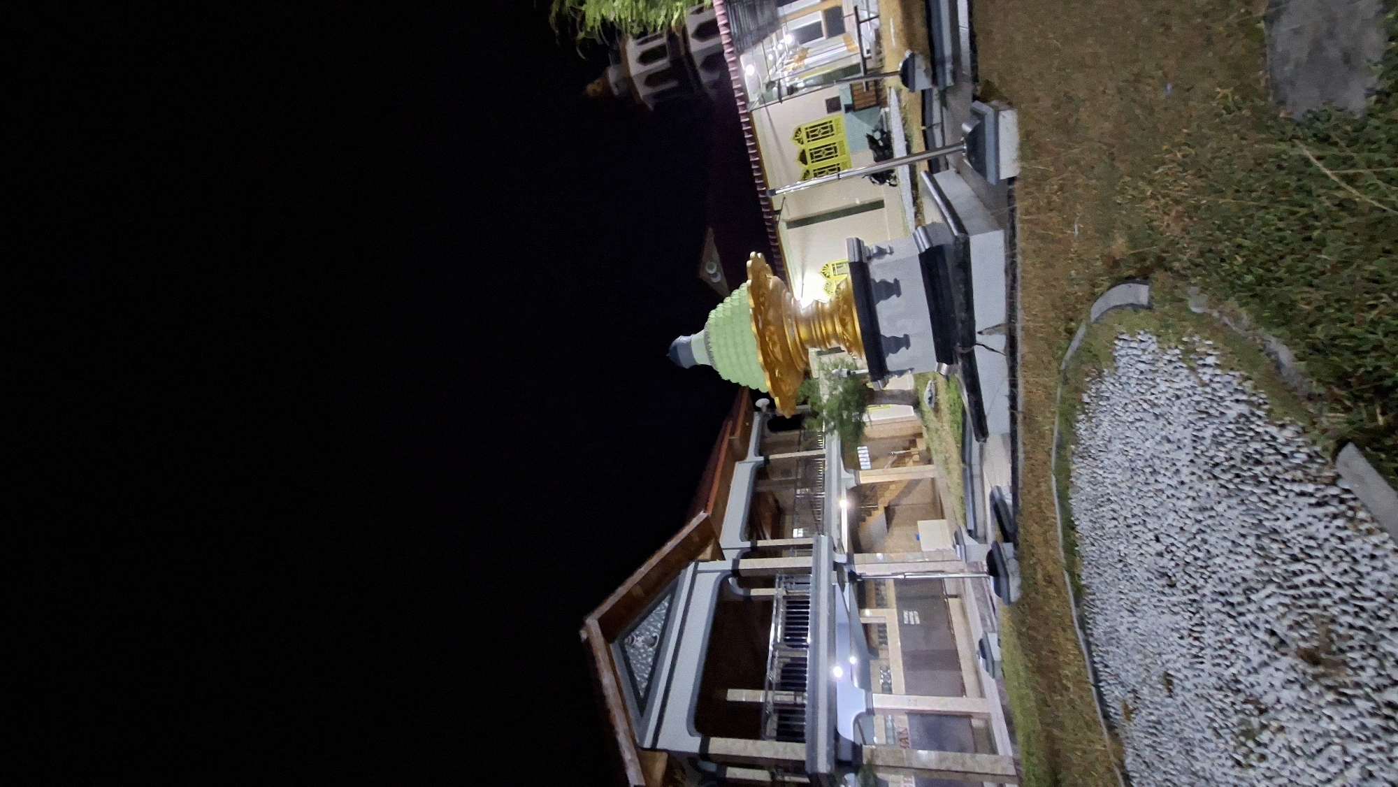 A nighttime shot captures a building with a layered roof and balconies, suggesting it is a mosque or a similar religious structure in Lhokdagang, Indonesia. In front of the building, a prominent gold and green tiered monument stands on a dark base. To the right, a minaret with white arches and a dome is visible, further confirming the religious context. The foreground features a circular area filled with small white pebbles, surrounded by dry grass. The overall scene is dimly lit by artificial lights, creating a contrast between the illuminated structures and the dark night sky.
