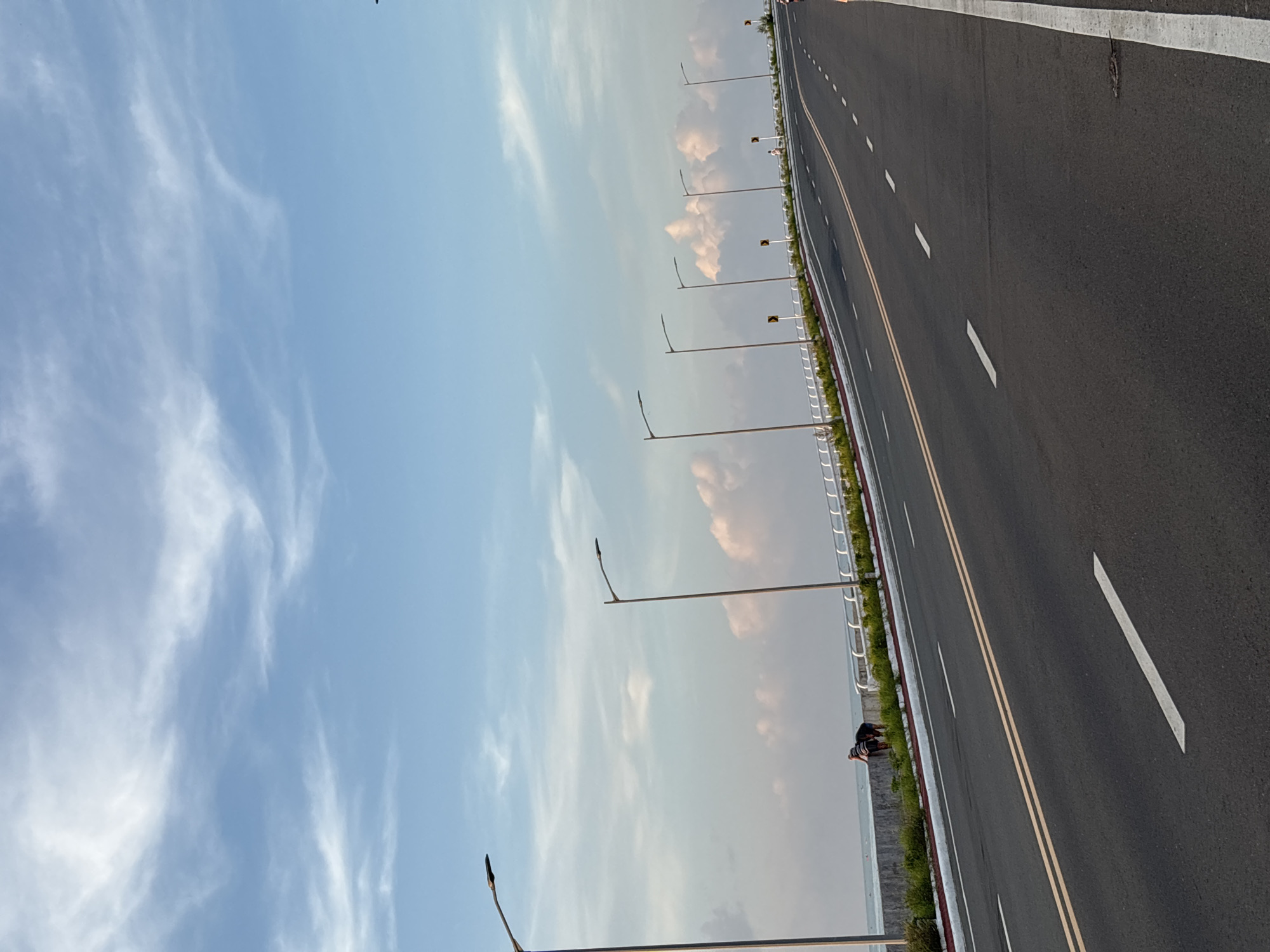 The image depicts a scenic view of a coastal highway in Davao City, Philippines, likely at dawn or dusk, given the soft lighting and pastel hues of the sky. The highway stretches into the distance, its asphalt surface marked with double yellow lines and dashed white lines indicating lanes. To the left of the road, a concrete barrier separates it from a body of water, presumably the sea, with a white railing running along the top. A line of modern streetlights stands at regular intervals along this barrier, illuminating the pathway. Two individuals are visible near the barrier, one bending down, their actions unclear but suggesting interaction with the environment. Beyond the barrier and water, the sky is a canvas of pale blue with wispy white clouds and larger, billowy pinkish clouds, characteristic of a tropical sky. The overall impression is one of serenity and an expansive landscape.