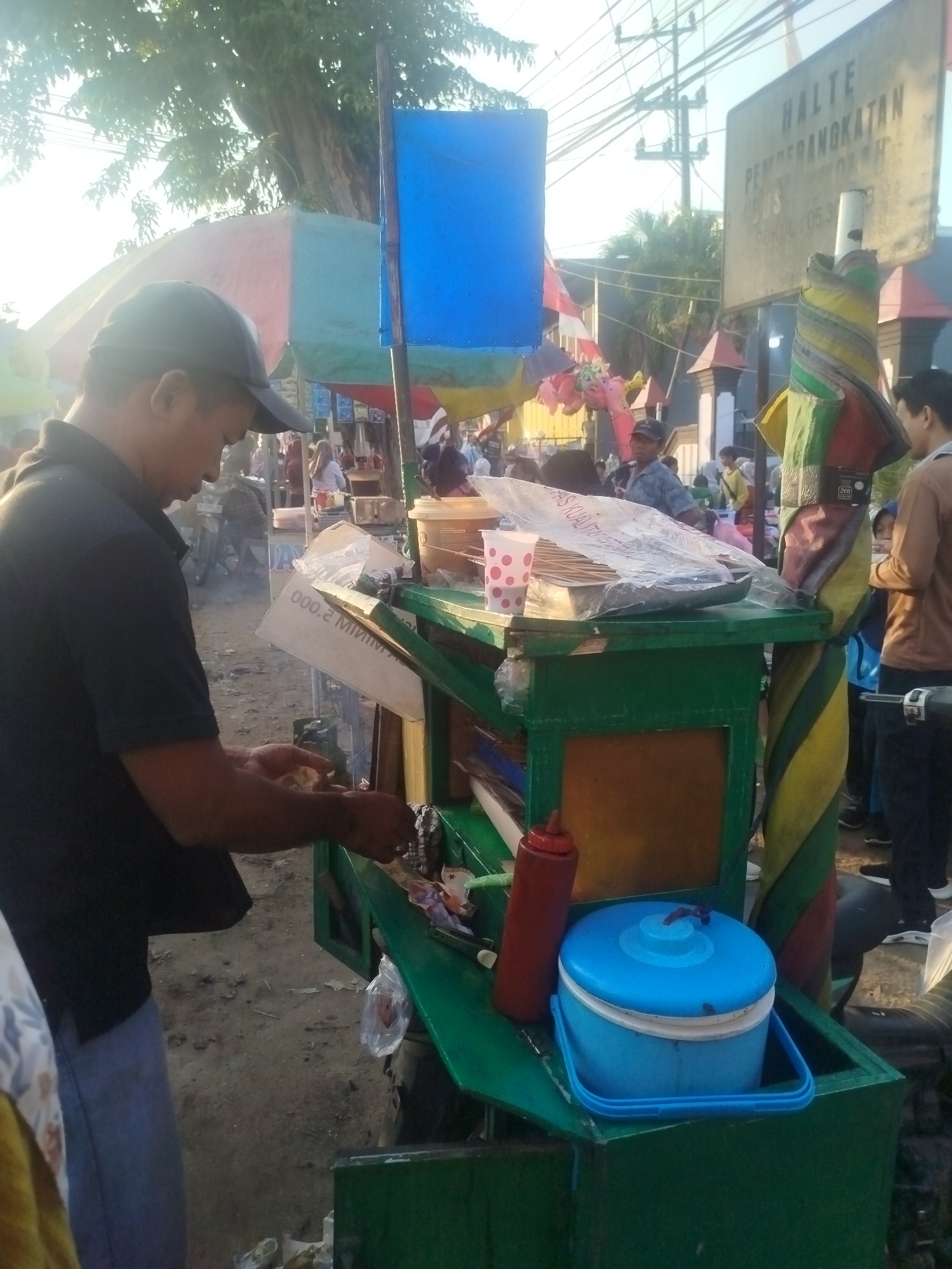 The image depicts a street scene in Burneh, Indonesia, characterized by an outdoor market or food stall area. A man wearing a black t-shirt and a dark baseball cap is the central human subject, actively engaged in preparing food at a green food cart. The cart itself is laden with various items, including containers of food, drinks, a red squeeze bottle, and a blue cooler. Overhead, colorful awnings and umbrellas provide shade. In the background, other people are visible, suggesting a bustling public space. A sign with text is mounted on a pole, indicating a "HALTE PEMBERANGKATAN" (Departure Stop) with times listed, implying a transportation hub or a busy intersection. The overall environment appears to be dusty and worn, consistent with an outdoor market setting. The lighting suggests it is daytime, possibly late afternoon given the warm tones.