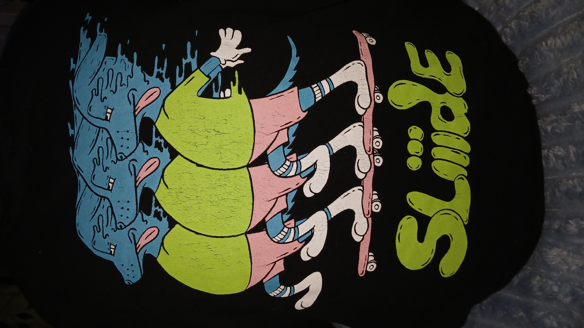 The image shows a black t-shirt with a colorful cartoon-style graphic printed on it. The graphic features a stacked repeating image of a cartoon character riding a skateboard. The character appears to have a blue dog head followed by a green shirt and pink pants. The text "Slide" is printed vertically in a stylized green font to the right of the character. The overall style is reminiscent of skate culture and pop art. There is no indication the image was taken in the city of Dnipro, Ukraine.
