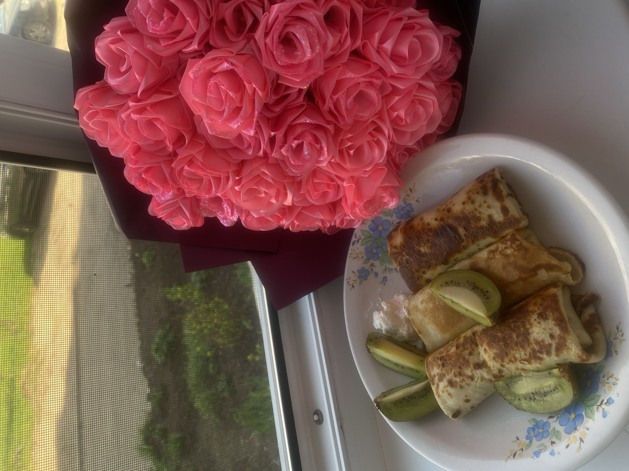 A bouquet of pink roses sits next to a white plate filled with folded crepes, sliced kiwi fruit, and a dollop of cream. The objects are positioned on a white windowsill next to a window with a screen. Outside the window, there is a partial view of vegetation. This image does not provide sufficient information to definitively determine the location as Chaikovsky, Russia, or to identify any individuals or interactions.