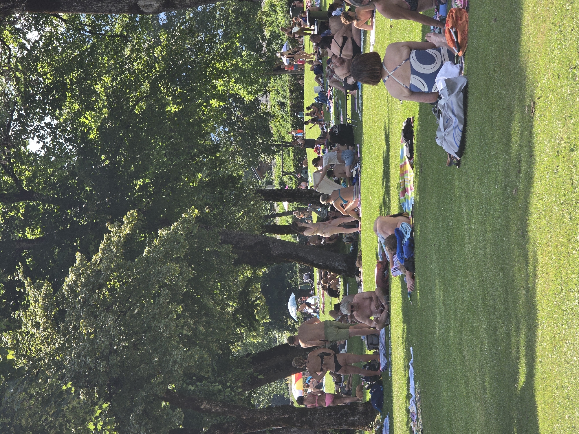 This media file depicts a vibrant scene of people enjoying a sunny day on a grassy lawn, likely in Garmisch-Partenkirchen, Germany. The image is oriented vertically, with the lush green grass and scattered trees dominating the frame. A significant number of individuals, dressed in swimwear, are spread across the lawn, sunbathing, socializing, and relaxing.
The activities suggest a warm day, possibly a summer afternoon, given the bright sunlight casting distinct shadows and the attire of the people. The presence of towels, blankets, and bags indicates a leisure outing, possibly at a park, a beach, or an outdoor recreational area. Some individuals are lying down, while others are seated or in casual conversation. The overall atmosphere appears relaxed and carefree.
Notable details include the vibrant green of the well-maintained grass and the dappled sunlight filtering through the dense foliage of the trees. The shadows suggest the sun is moderately high in the sky. The people exhibit a range of body types and ages, contributing to a diverse and inclusive gathering. While no specific text is clearly legible, the overall impression is one of community and enjoyment of the outdoors. The scene evokes a sense of summer relaxation and a peaceful escape from daily routines.