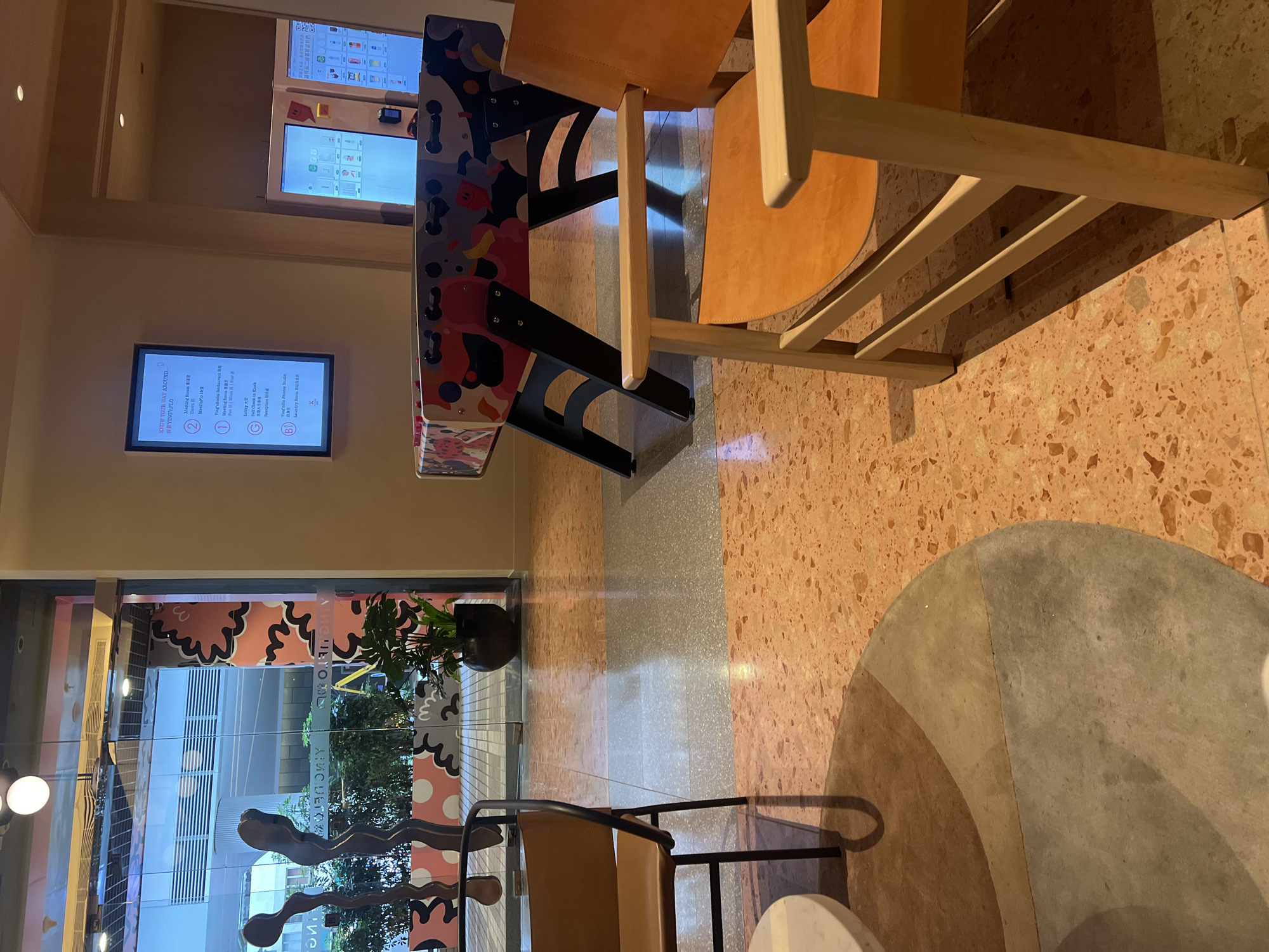 This image depicts the interior of a modern co-working space in Hong Kong, China. The scene features a variety of furniture and amenities, suggesting a lively and collaborative environment.
In the foreground, a light wood-framed chair with an orange seat is positioned on a textured floor. To the left, another chair with a brown seat and black frame is visible, situated near a round, light-colored table. The floor itself is a striking mosaic of pinkish-orange terrazzo with grey and beige circular patterns.
Dominating the mid-ground is a colorful foosball table with a vibrant, graphic design. Beyond this, a digital display screen mounted on the wall lists various locations within the building, indicated by numbers and letters, and in both English and Chinese. Examples include "Meeting Room," "Ying's Social Restaurant," "Lobby," and "Laundry Room."
To the left, large glass doors offer a view into an outdoor or adjacent area, revealing greenery and a glimpse of urban architecture. Two abstract, dark sculptures are positioned near the entrance, adding an artistic element.
In the background, two illuminated vending machines, likely offering drinks or snacks, are visible against a plain wall. The overall ambiance is contemporary, functional, and designed to foster interaction and productivity. The presence of the foosball table, digital directory, and comfortable seating suggests a space that balances work with leisure and community building. The lighting appears to be a mix of ambient and focused, creating a warm and inviting atmosphere.