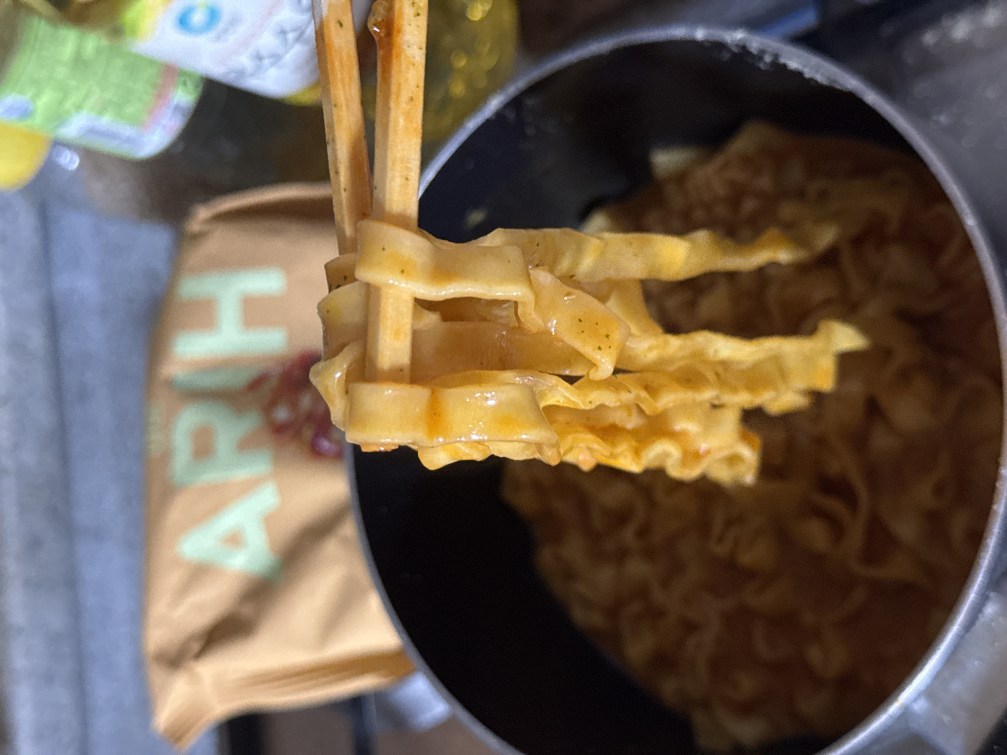 A close-up shot captures a hand holding chopsticks with freshly cooked, vibrant orange noodles, partially submerged in a pot of the same dish. The noodles appear to be wide and flat, with a slightly rippled texture, coated in a rich, spicy sauce. In the background, a blurred brown package with light green lettering, possibly containing the noodles, is visible along with two out-of-focus bottles. The lighting is natural, suggesting an indoor setting, possibly a kitchen, given the presence of a pot and bottles. The image is static and does not depict any action or interaction between people.