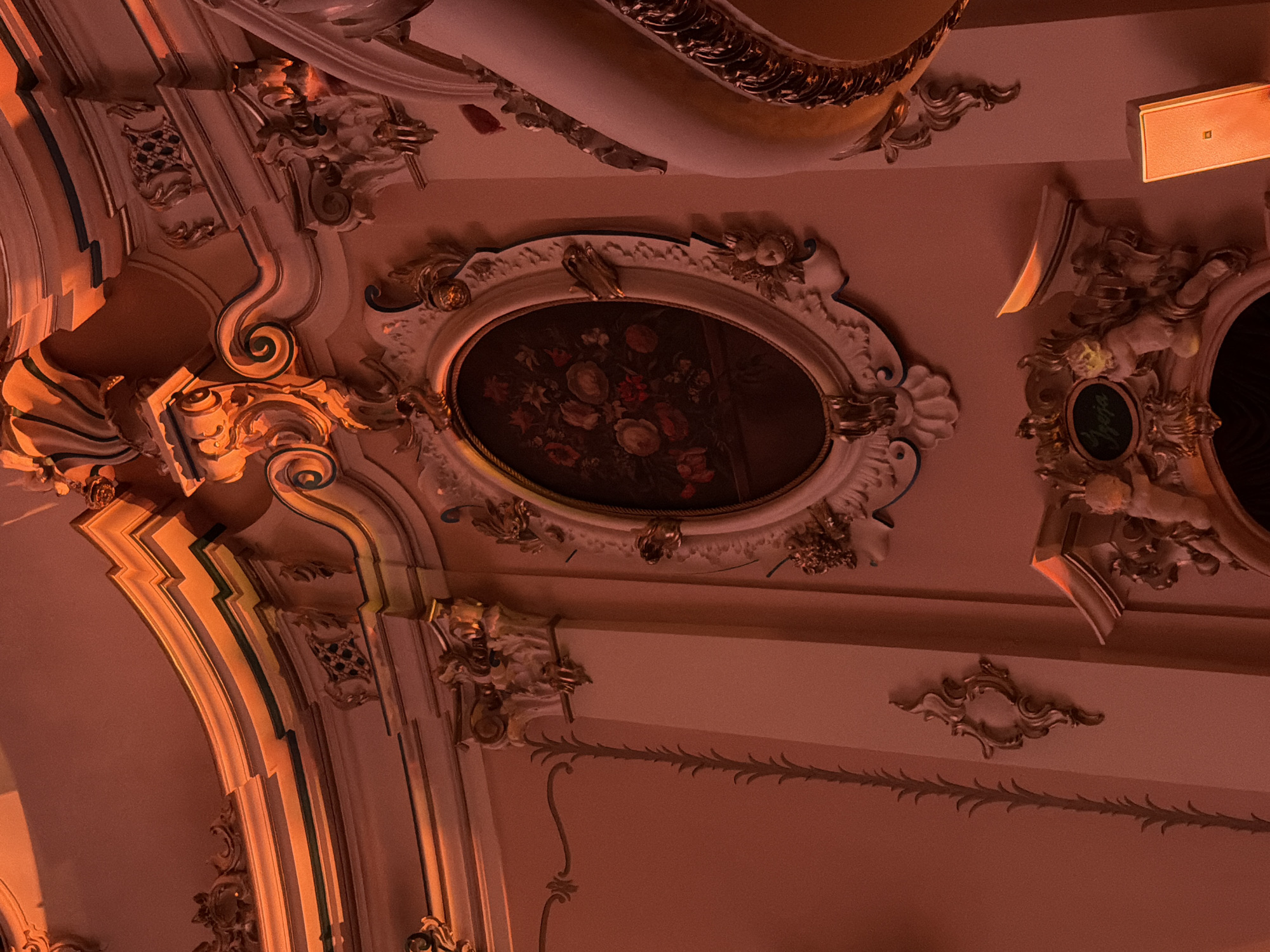 The image depicts the ornate interior of the Riga Russian Drama Theatre in Riga, Latvia. The scene is bathed in warm, orange lighting, highlighting the intricate architectural details. A central oval-shaped painting, featuring a floral arrangement, is framed by elaborate gilded mouldings. The walls are adorned with decorative plasterwork, columns, and sculpted elements, including cherubs near the bottom of the frame. The overall impression is one of grandeur and historical richness characteristic of theatre interiors from the late 19th or early 20th century.
