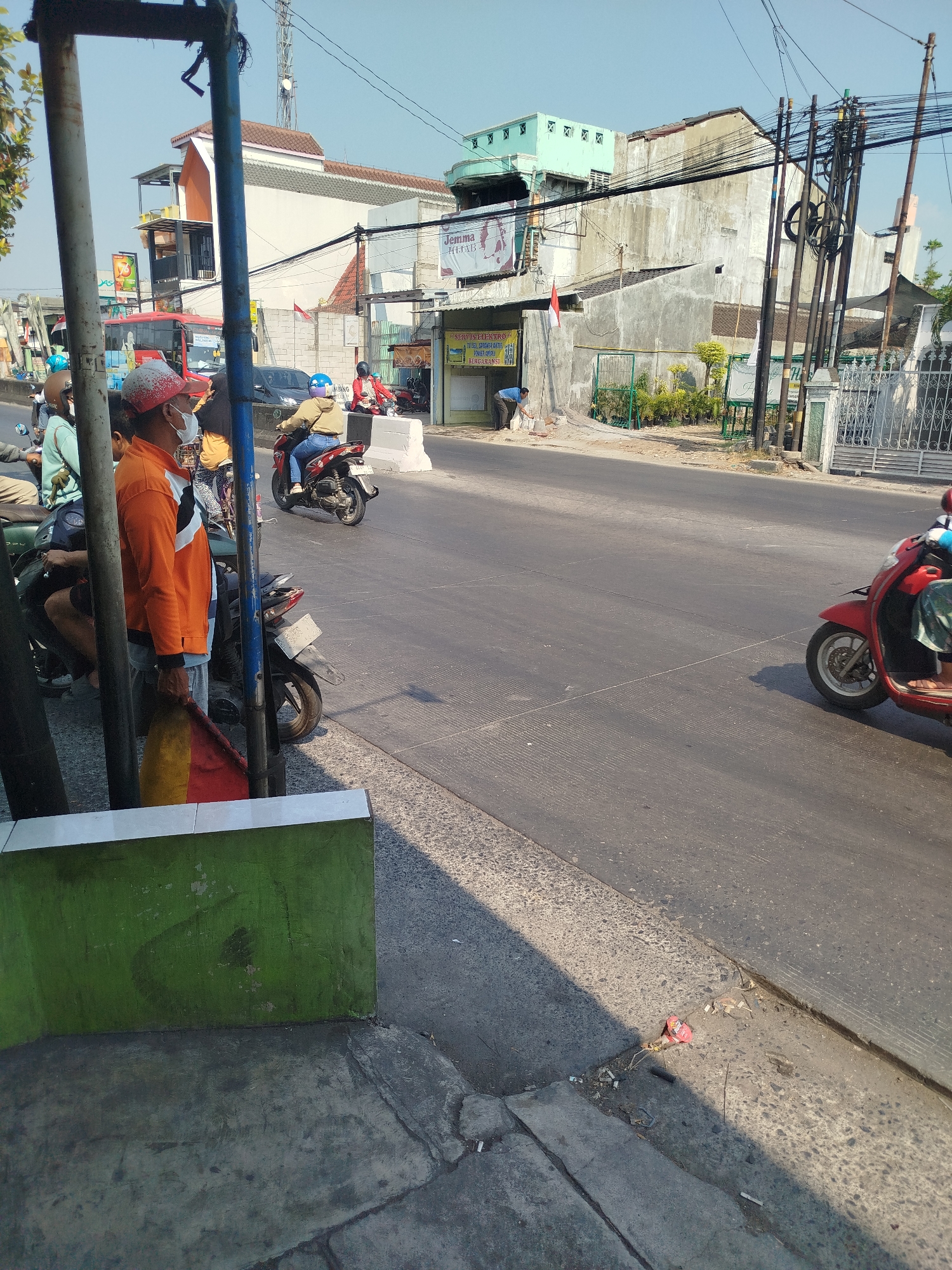 In Mranggen, Indonesia, a street scene unfolds under a clear sky. On the left, a person wearing an orange jacket and a face mask stands near a metal pole, holding a red and yellow flag. They are accompanied by several motorcycles parked nearby, with riders in helmets. The street is paved, with a sidewalk on the left lined by a low green wall. In the background, buildings of varying sizes and architectural styles line the street, some appearing to be commercial establishments with signage. The overall environment suggests a typical urban or semi-urban setting with ongoing street activity.