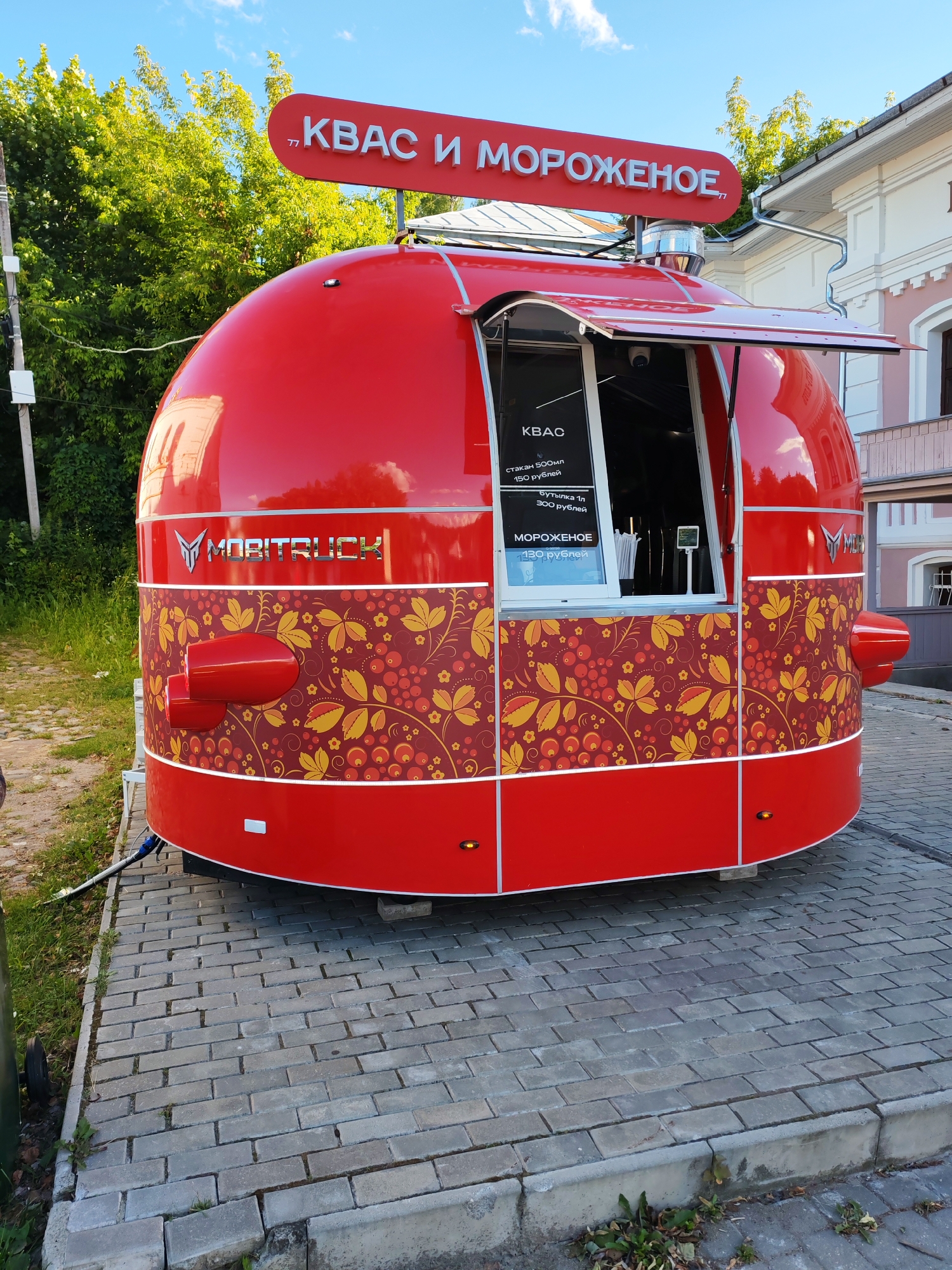 This is a vibrant, eye-level photograph of a red mobile food truck, likely selling kvass and ice cream, parked on a brick-paved area. The truck has a distinctive, rounded, almost pod-like shape, with a bright red exterior adorned with a decorative pattern of golden leaves and red berries. Above the serving window, a prominent red sign in white lettering reads "КВАС И МОРОЖЕНОЕ," meaning "Kvass and Ice Cream" in Russian.
Inside the open serving window, a menu is visible, listing prices for kvass (500ml for 150 rubles, 1-liter bottle for 300 rubles) and ice cream (130 rubles). The truck also features the "MOBITRUCK" logo on its side, a stylized winged emblem. Two red, cylindrical elements, possibly exhaust vents or decorative features, are mounted on the lower sides of the truck.
The scene suggests it's a bright, sunny day, indicated by the clear blue sky with scattered clouds and the overall well-lit environment. Lush green trees are visible in the background, and a light-colored building with architectural details can be seen to the right. The truck is positioned on a pathway made of rectangular grey bricks, with some grass and weeds growing along the edges, hinting at an outdoor setting, possibly in a town or park. There are no people visible in the frame, and the overall atmosphere is clean and inviting, promoting the sale of beverages and frozen treats. The location context provided is Plyos, Russia.