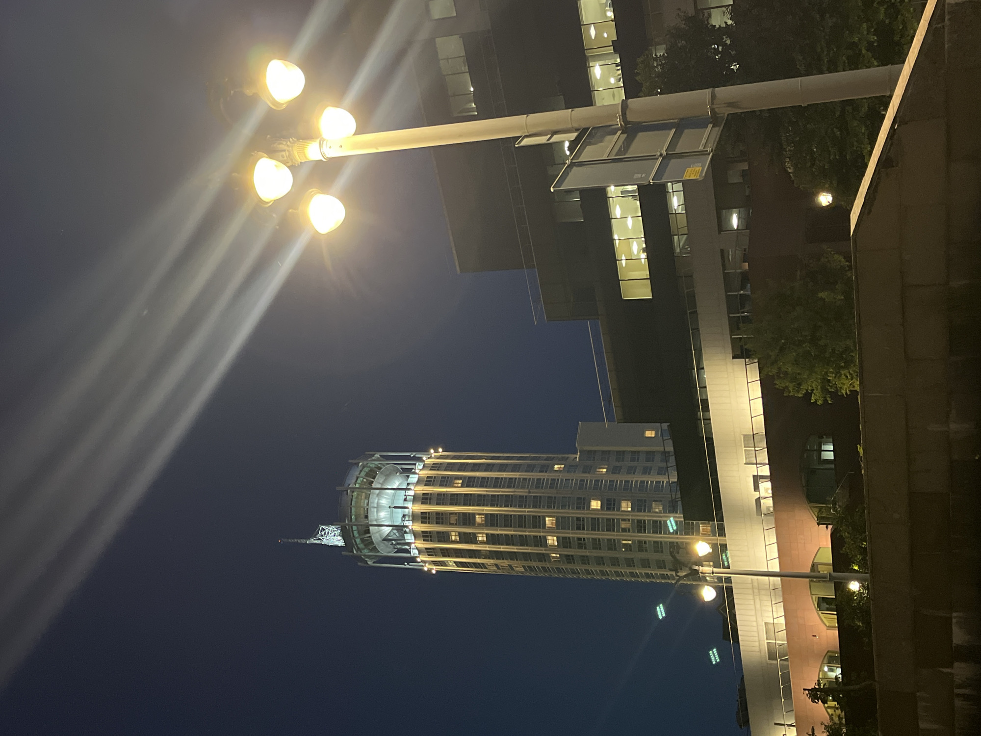 The image depicts a nighttime view of the building complex at 18 Bolshaya Yakimanka Street in Moscow, Russia. The prominent structure is a tall, cylindrical skyscraper with a circular, illuminated crown, topped by a radio mast. The skyscraper's facade is characterized by numerous vertical rows of windows, many of which are lit, indicating occupancy. Adjacent to the skyscraper is a lower, darker, multi-story building with large windows that also display interior lighting.
In the foreground, a lamppost with four glowing bulbs stands prominently on the right side of the frame. The light from these bulbs creates streaks across the dark night sky. Below the lamppost and extending to the left, there is a raised stone embankment or wall, partially obscuring the lower levels of the building complex. Trees with foliage are visible behind the embankment and around the base of the buildings. The sky is a deep blue, with faint cloud-like streaks illuminated by light sources, possibly from searchlights or atmospheric conditions. There are no people or vehicles visible in the image.