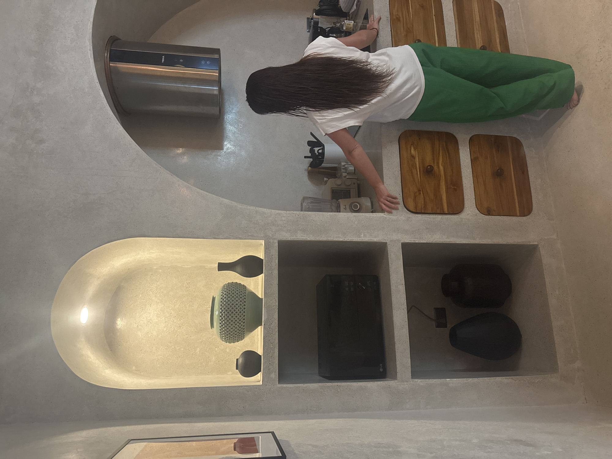 An interior scene depicts a modern kitchen or kitchenette area located in El Nido, Philippines. The walls and integrated shelving are constructed from a light grey, textured material resembling polished concrete, featuring prominent arched recesses.
One individual, a person with long, dark, wet hair, is visible from behind, positioned at a light-colored countertop. They wear a white short-sleeved t-shirt and loose-fitting green trousers, and appear to be barefoot. Both hands rest on the countertop, suggesting a pause or preparation.
To the left, a built-in shelving unit with three levels is present. The top level, an arched niche illuminated by a recessed light, displays three decorative vases: a large, textured, light teal vase centrally flanked by two smaller, dark, simple vases. The middle shelf holds a black LG microwave oven. The bottom shelf contains two dark, distinct decorative objects, one teardrop-shaped and one cylindrical. A partially visible framed artwork hangs on the far left wall.
Above the countertop, a stainless steel cylindrical range hood branded "TEKA" is mounted within an arched recess. The countertop itself holds various kitchen items, including a light-colored blender, a white utensil holder with kitchen tools, and a small bottle among other indistinct objects. Below the countertop, three wooden cabinet doors with rounded corners and circular knobs are visible, suggesting storage. The floor matches the light, textured aesthetic of the walls.