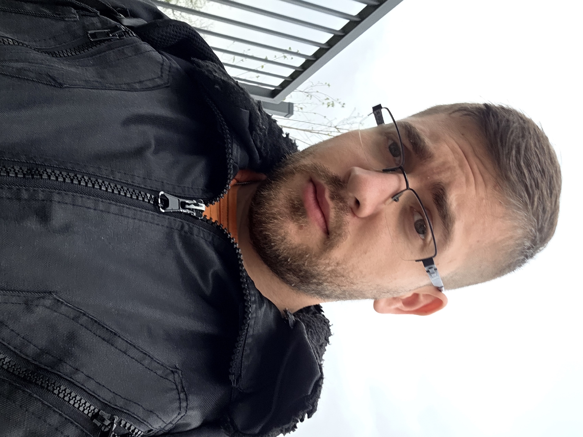 A man with short brown hair, a beard, and glasses poses for a photo while standing underneath a metal structure, possibly a pergola or awning. He is wearing a black zippered jacket over an orange shirt. The background is blurry and suggests an outdoor setting, although specific landmarks of Plovdiv, Bulgaria, are not discernible.