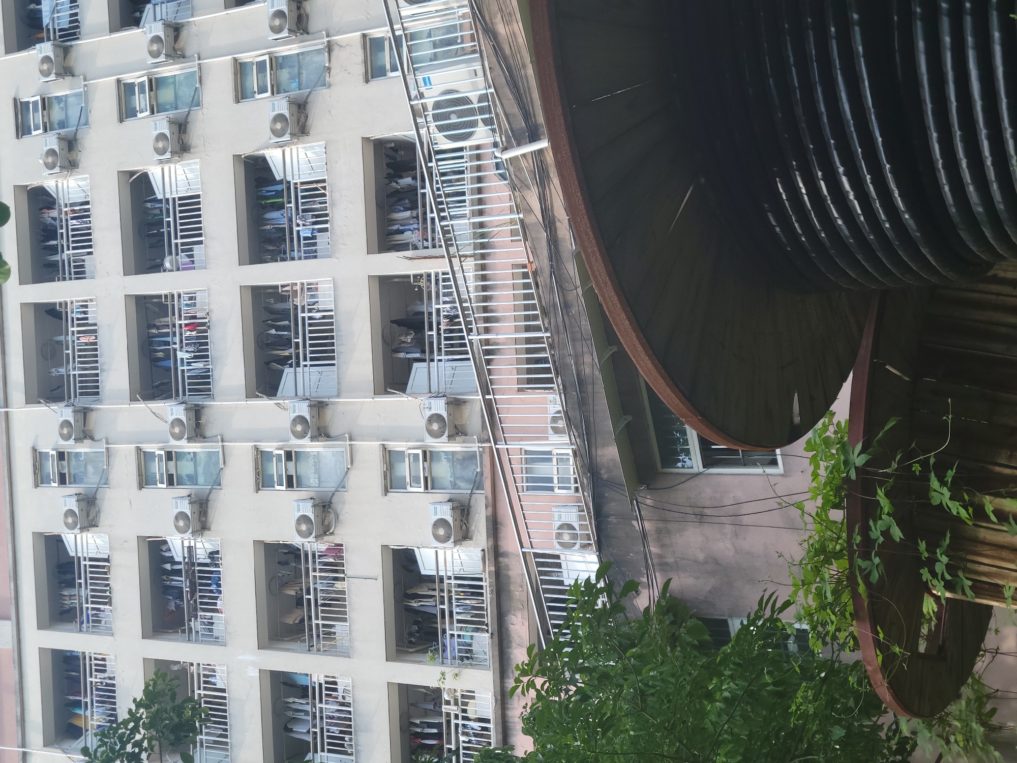 A rotated view of an apartment building in Beibei, China, showing numerous windows, each equipped with an air conditioning unit and safety bars. Clothes are drying on racks inside some of the windows. A dark, curved, metallic structure is situated to the right of the building, partially obscured by foliage. Utility lines run along the side of the building.