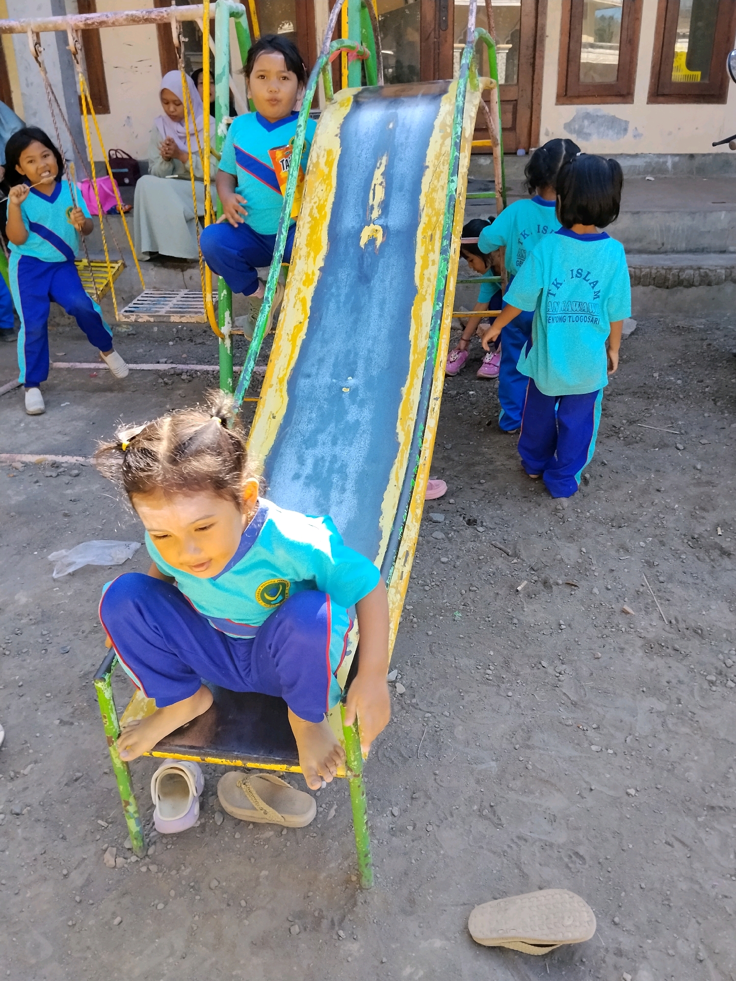 In Tlogosari, Indonesia, on a dirt ground, a group of young children are playing on a colorful, worn playground. A metal swing set with yellow chains is visible on the left, with two girls in blue and teal uniforms on it. One girl swings, while the other, in a teal shirt with blue pants, chews on a stick. Behind them, a woman in a pale headscarf and long dress sits on a step, looking downwards. A third girl in a similar uniform stands on the swing set's platform, facing the camera with a surprised expression. In the center, a bright yellow and blue slide with worn sections and a green metal frame is the focal point. A young girl with braided pigtails, her face powdered with yellow makeup, sits at the bottom of the slide, looking down. She wears a teal shirt with blue trim and blue pants. To the right of the slide, two children in teal shirts and blue pants are positioned. One, facing away, appears to be about to descend the slide, while the other stands near the bottom. The overall environment is dusty and sunlit, with a building visible in the background.